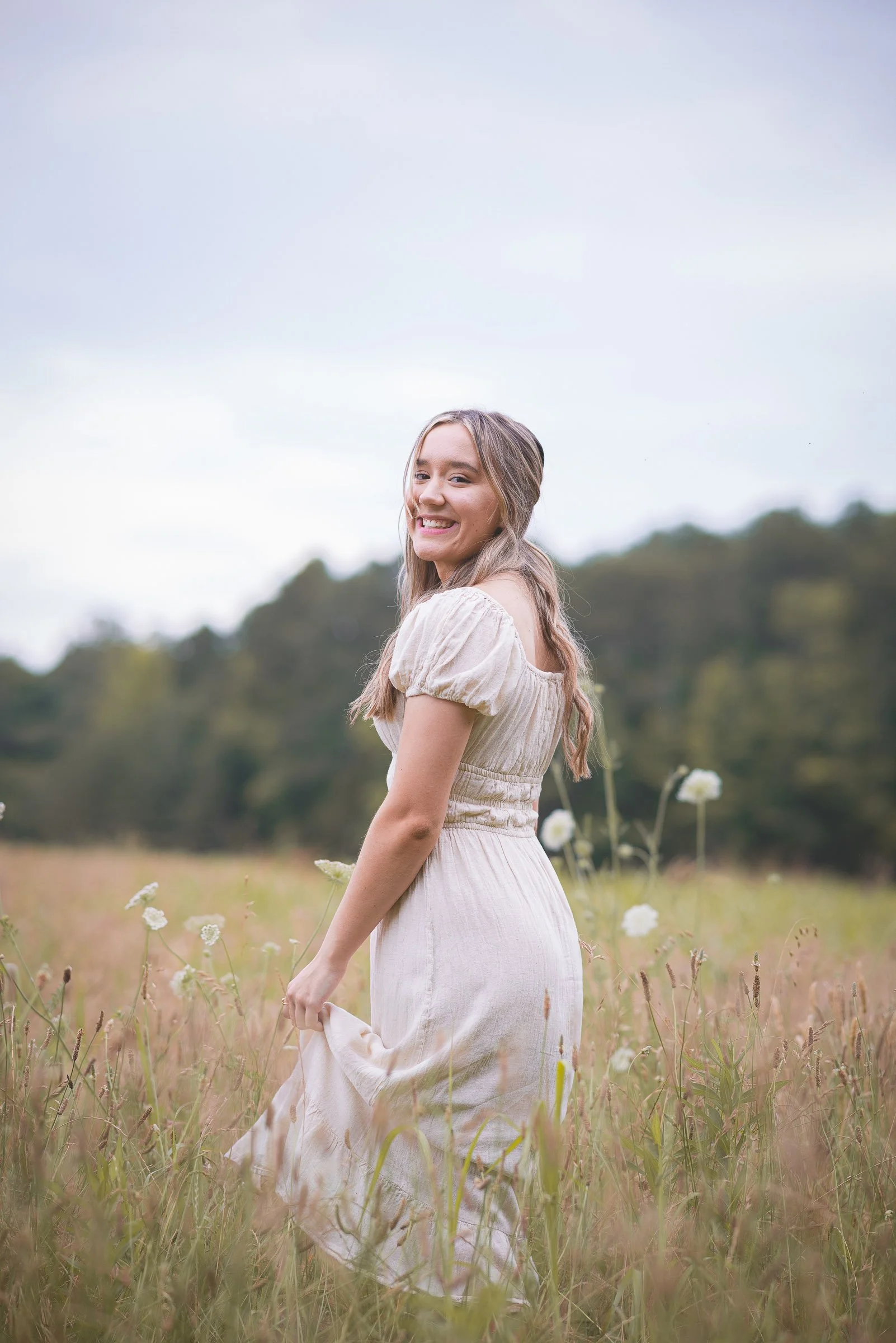 A young woman with long hair in a white dress standing in a grassy field with wildflowers, smiling at the camera, with trees and a cloudy sky in the background.