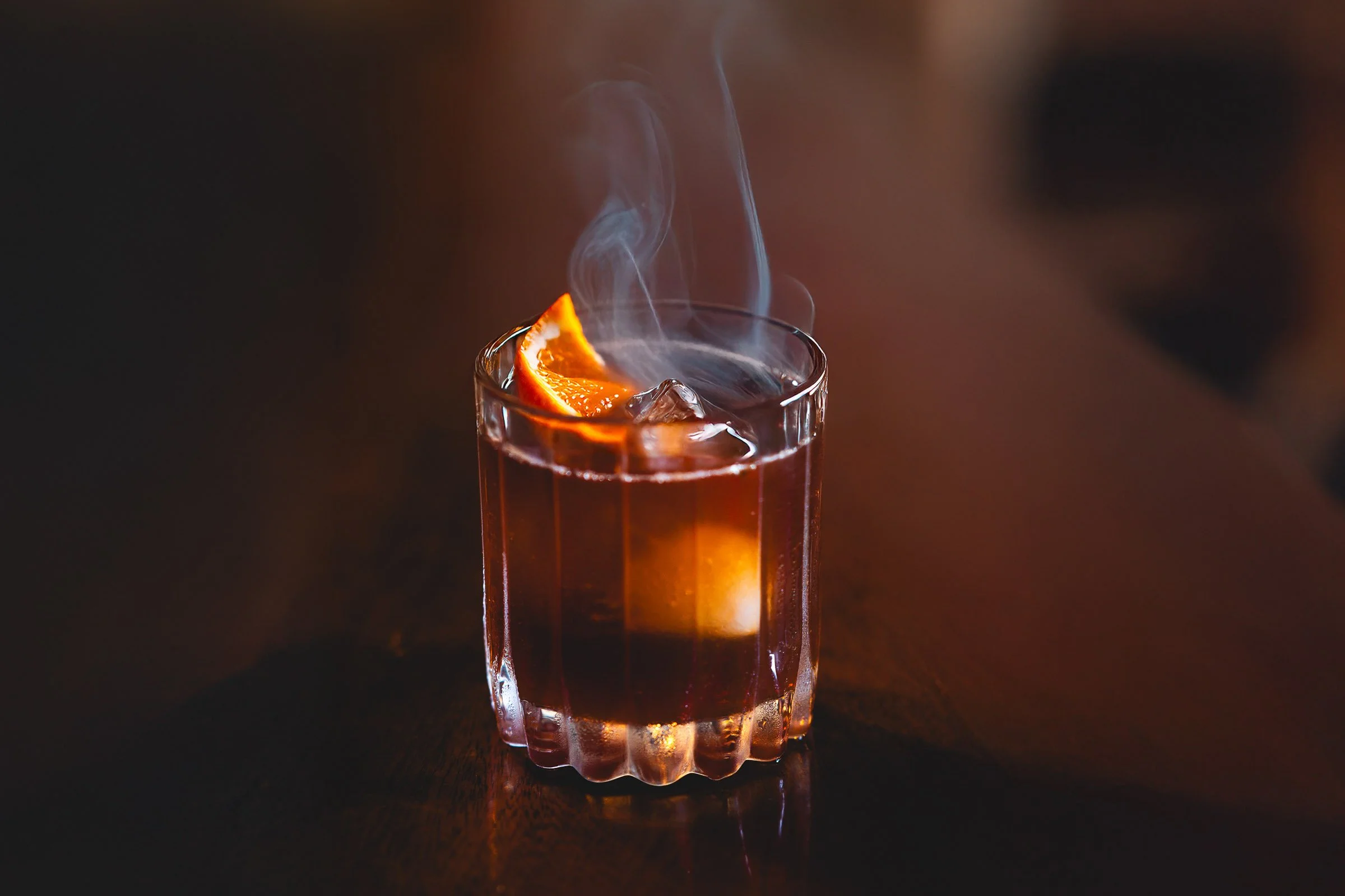 A glass of hot mulled wine with an orange slice and ice cubes, emitting steam, on a dark wooden surface.