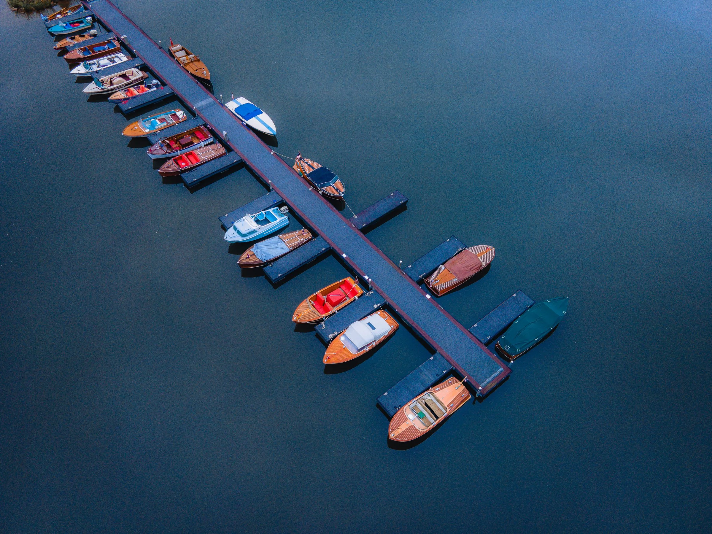 An aerial view of a dock with boats moored on both sides, floating on a calm dark water body.