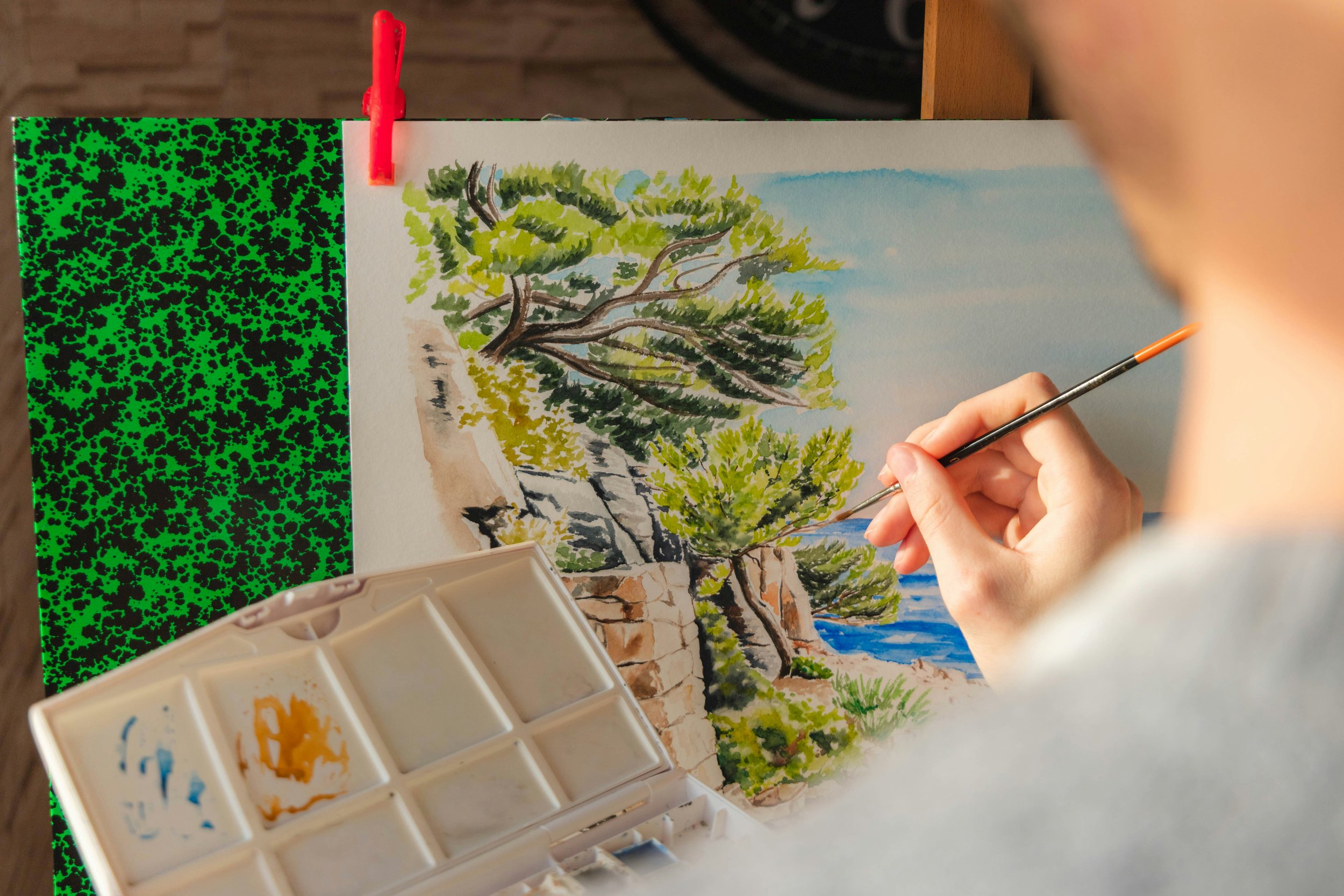 Main peignant un paysage de bord de mer à l’aquarelle, avec une palette de couleurs à côté.