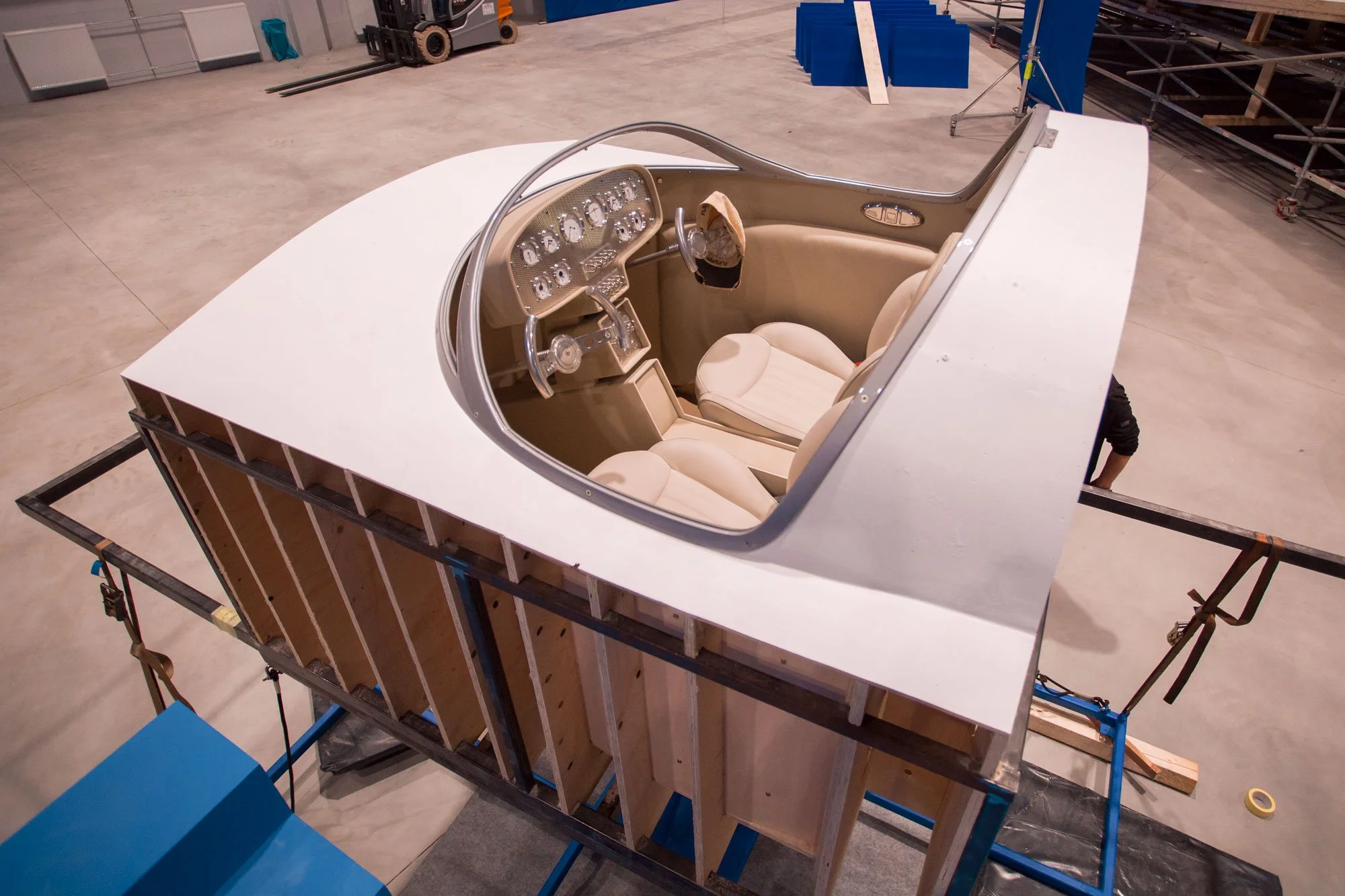 A car cockpit model being assembled inside a warehouse with a white outer shell, beige interior, and dashboard with gauges, set on a wooden frame.