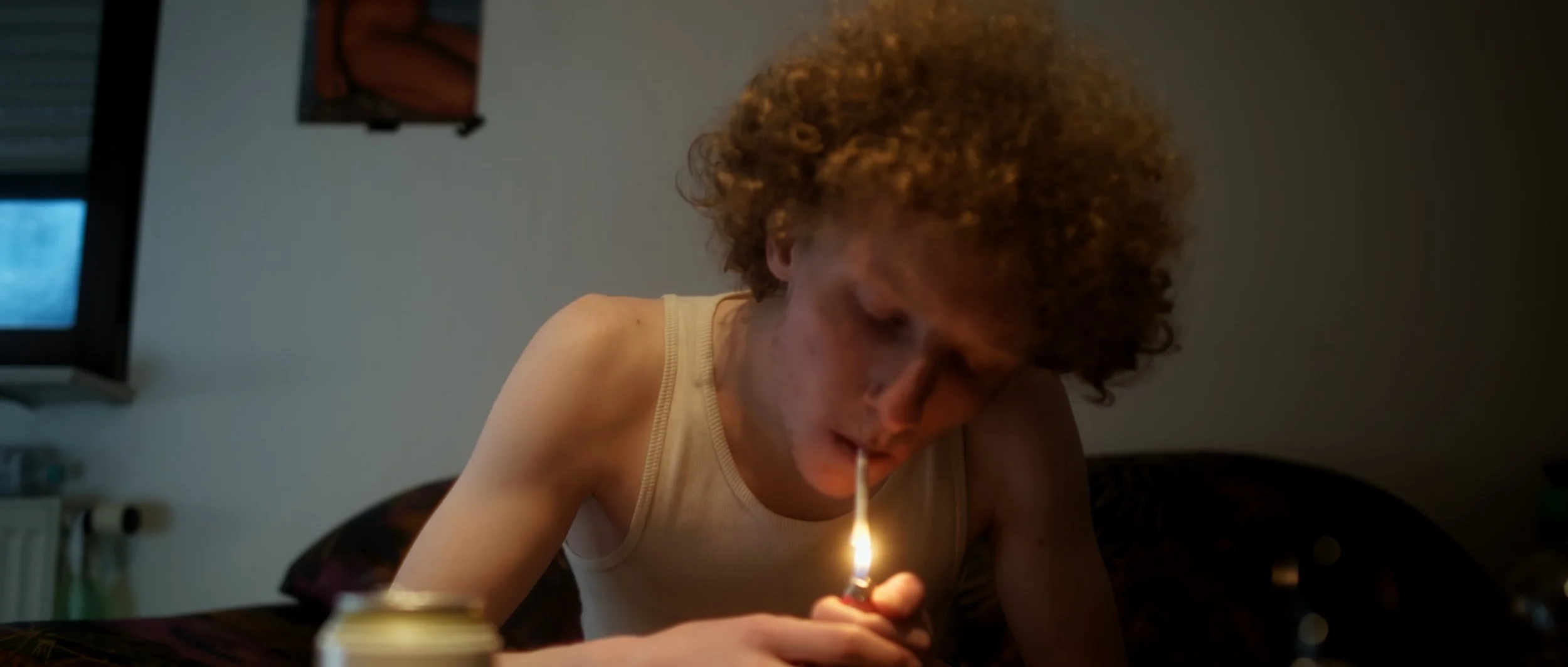 Junge mit curly Haaren in einem weißen Tanktop zündet eine Zigarette an.