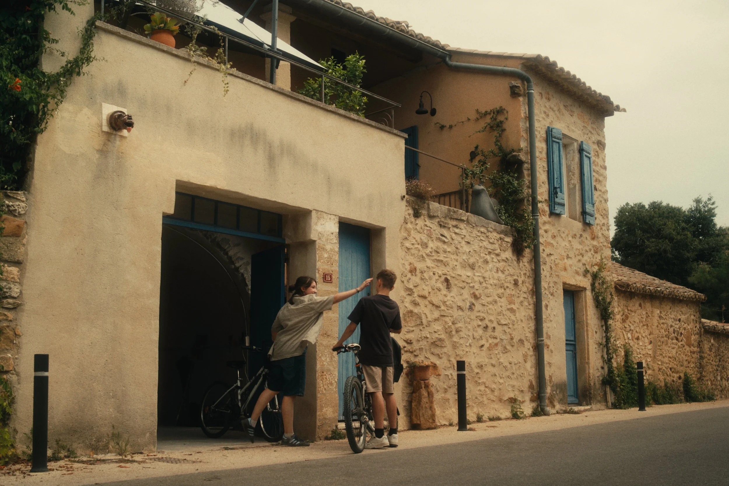 Zwei Jungen mit Fahrrädern stehen vor einem alten, Steinhaus mit blauen Fensterläden. Sie unterhalten sich, während eine von ihnen auf das Haus zeigt.