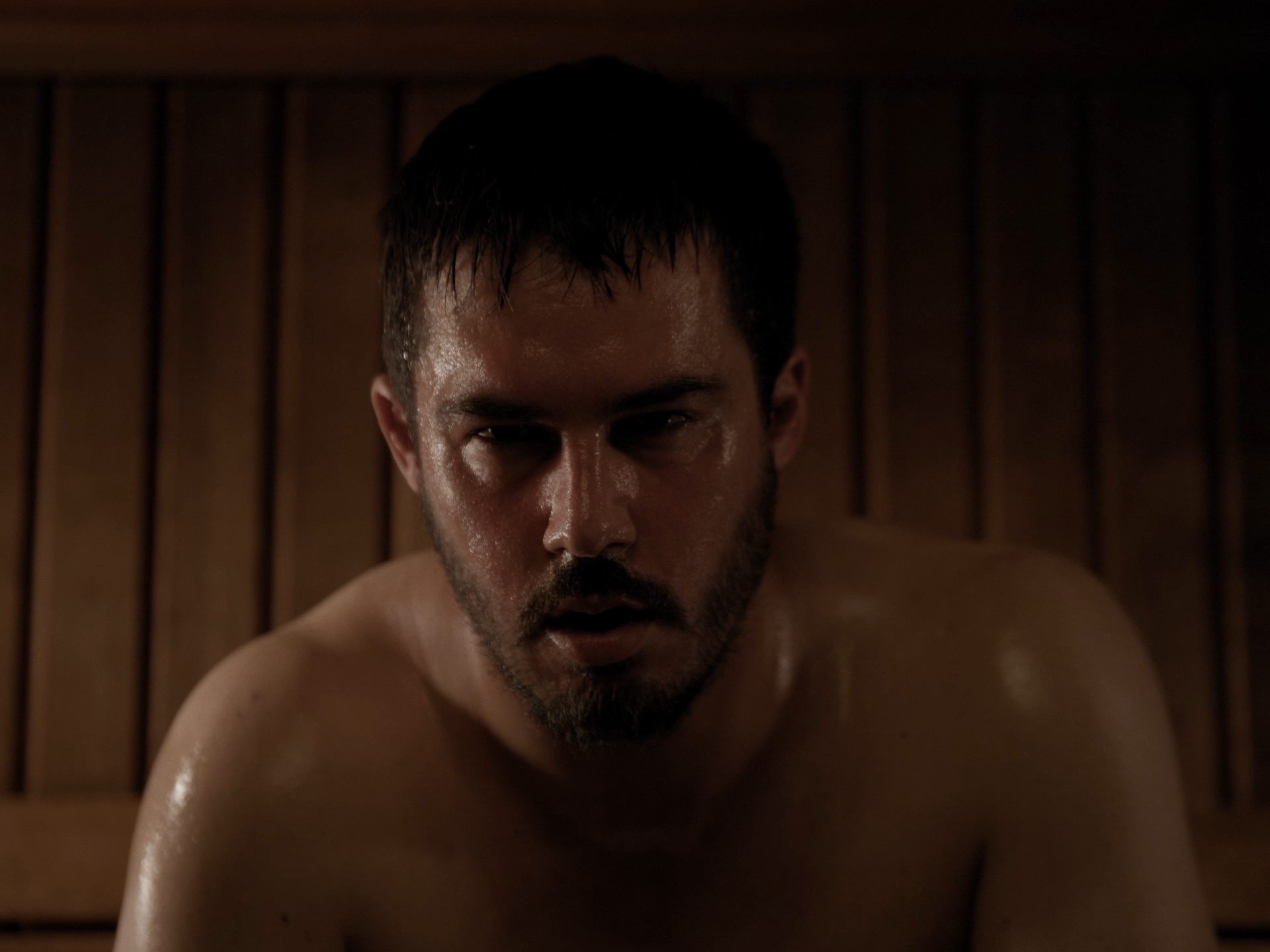 Ein Mann mit nassem Oberkörper sitzt in einem Raum mit dunklem Holz hintergrund, schaut ernst in die Kamera.