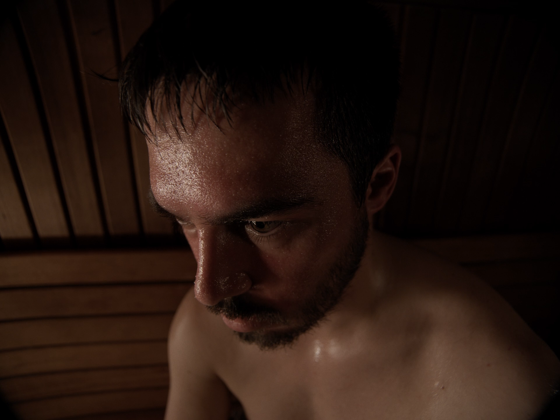 Ein junger Mann mit nassem Haar und leichtem Bart schaut nach unten in einem dunklen Raum mit Holzvertäfelung.