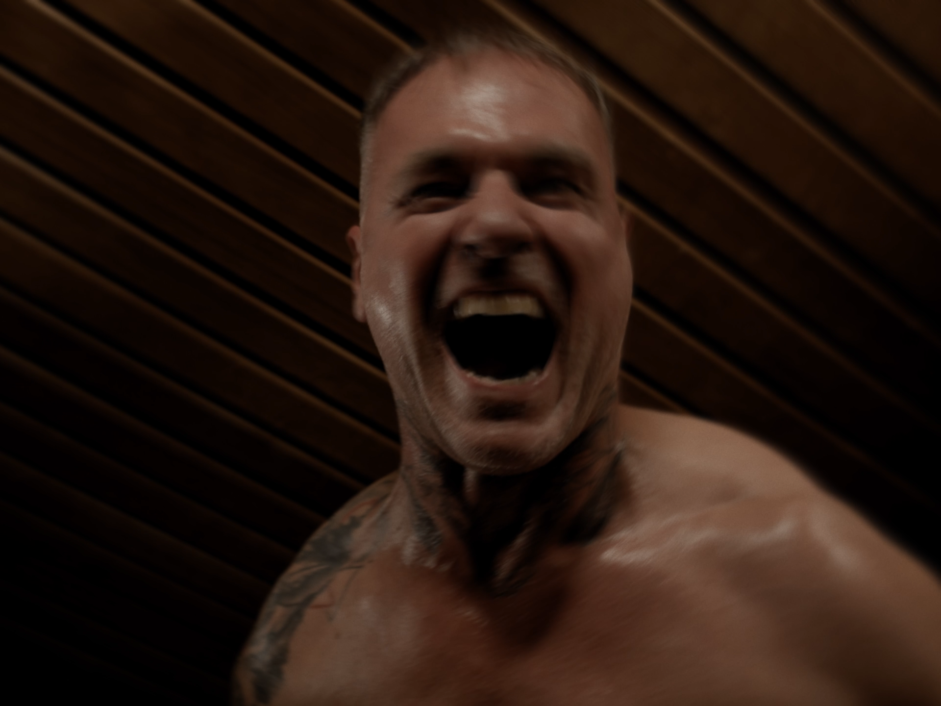 Ein Mann mit Tattoos auf der Schulter, nackt, schreit vor Wut, vor einer Holzdecke.