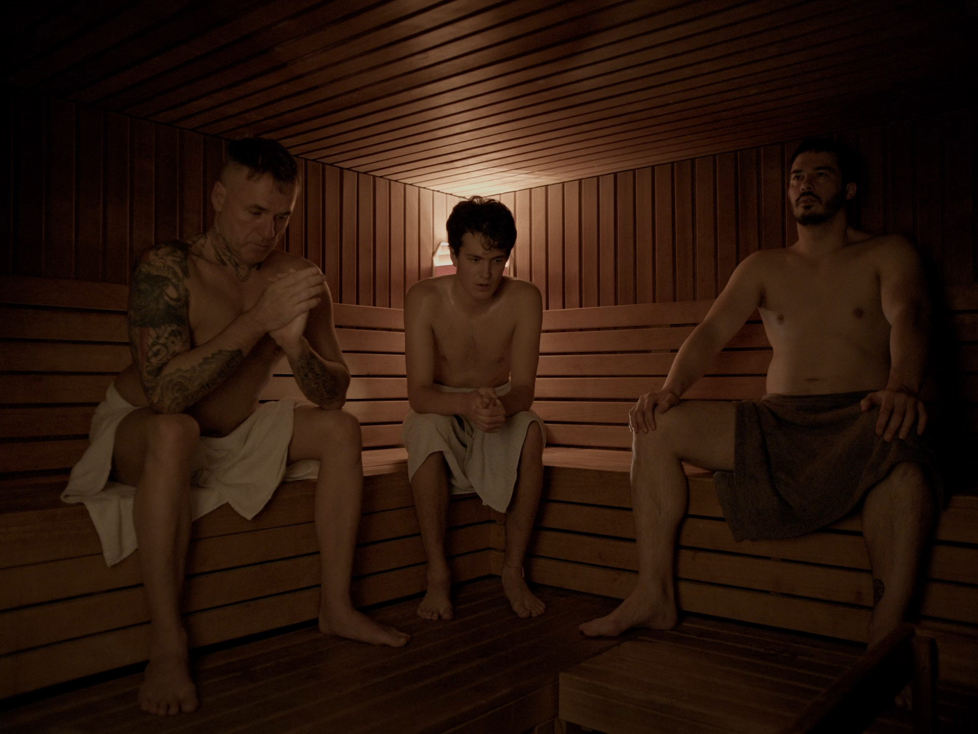 Drei Männer sitzen in einer Sauna, alle ohne Shirt, in entspannter Haltung.