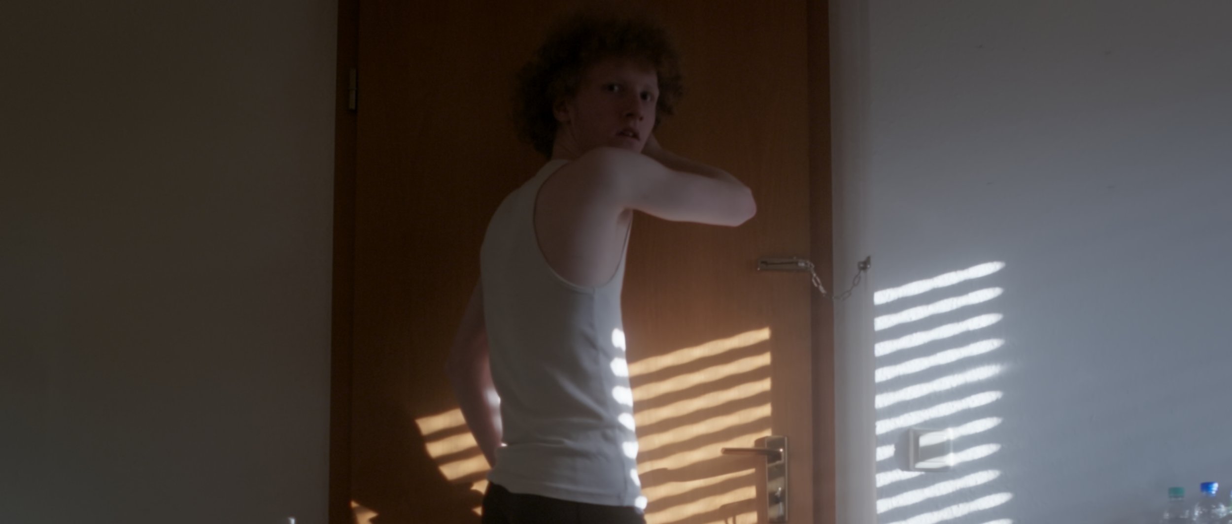 Junge mit lockigem Haar in einem weißen Tanktop steht vor einer Tür, wobei Lichtstrahlen durch Fensterläden auf die Wand neben ihm fallen.