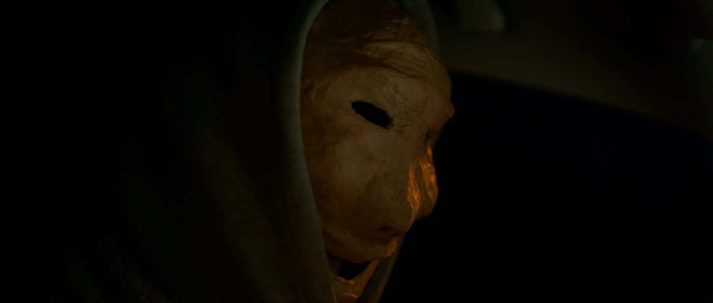 Ein maskierter Mann, nur teilweise sichtbar in dunkler Umgebung, blickt nach vorne.