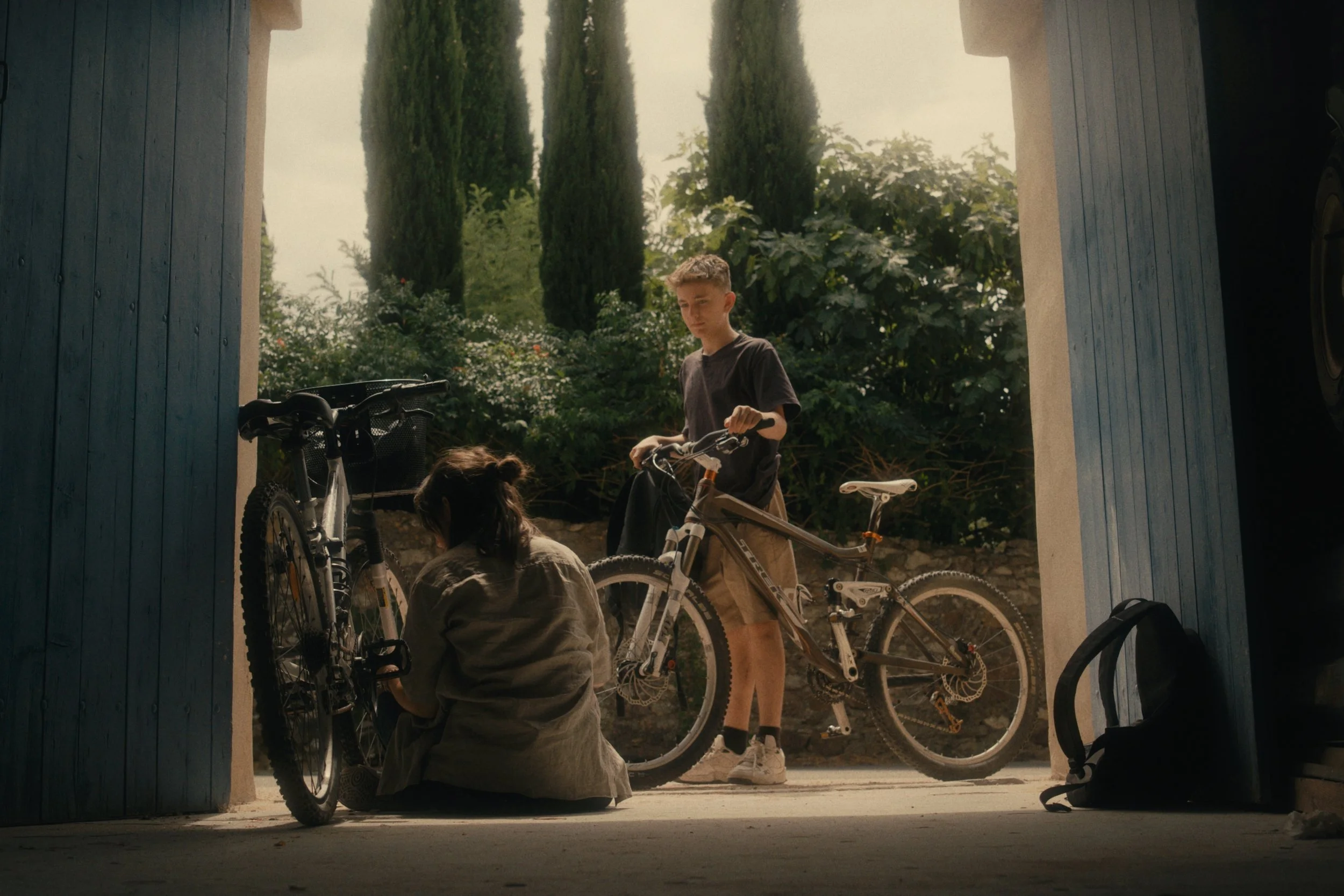 Ein Junge und ein Mädchen bereiten Fahrräder vor, im Hintergrund Bäume und Pflanzen, das Bild wird durch die Tür einer Garage betrachtet.