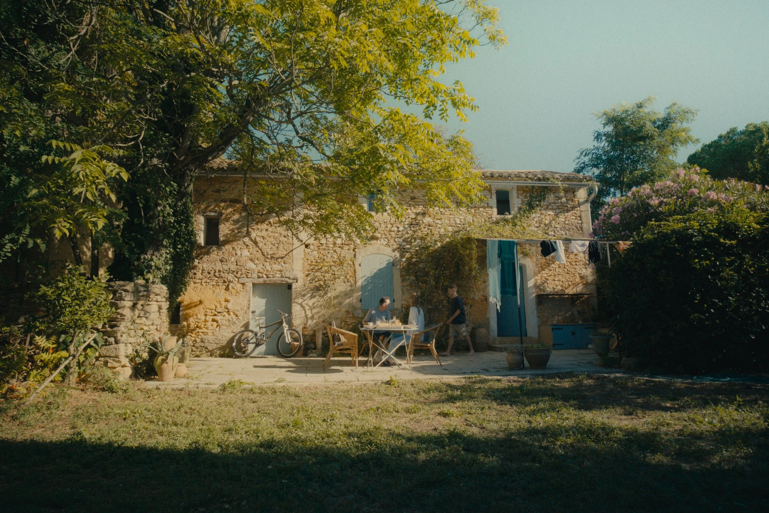 Ein rustikales Haus aus Stein mit blauer Tür und Fensterläden, umgeben von Bäumen und Blumen, mit einer Gruppe von Menschen, die im Garten sitzen und einen Tisch mit Essen und Getränken haben.