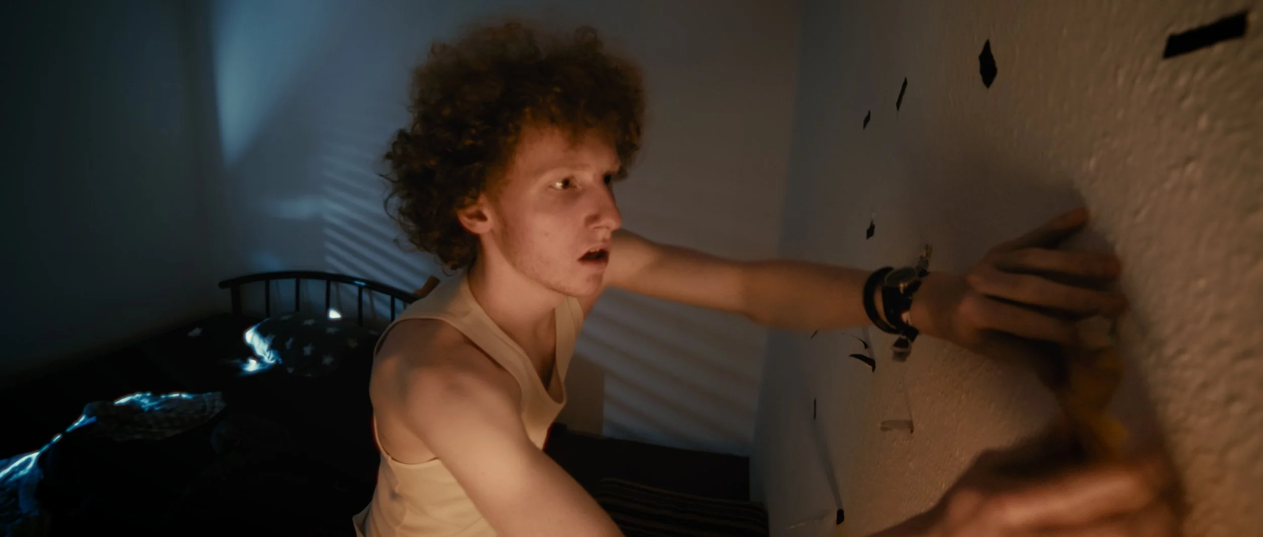 Junge Person mit lockigem Haar, lehnt an einer Wand und schaut überrascht oder erschrocken.