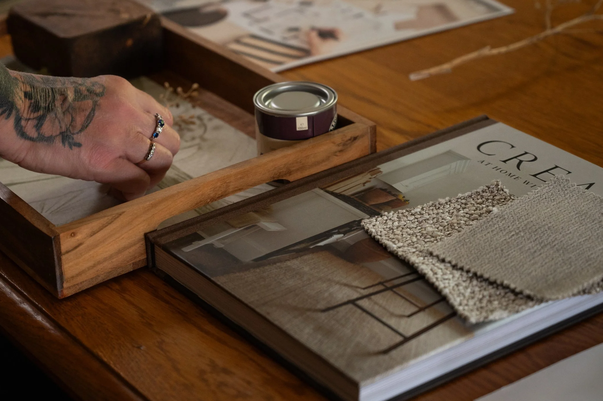 A person's hand with rings and tattoos reaching into a wooden box with a metal can inside, on a wooden table with magazines, a fabric sample, and decorative sticks nearby.