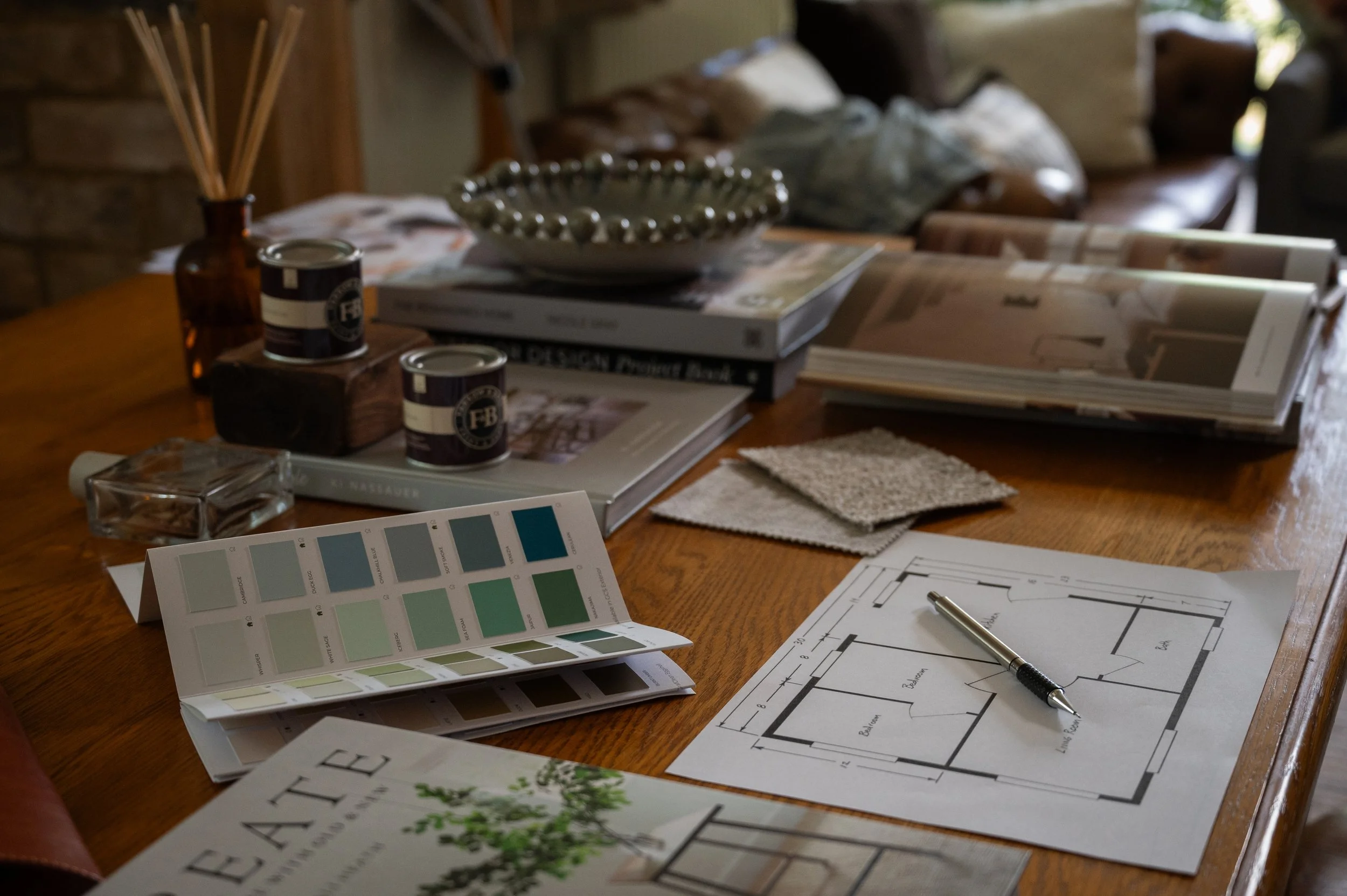 A wooden table with architectural plans, color swatches, magazine pages, pens, and samples, indicating interior design or home renovation planning in a cozy, well-lit room.
