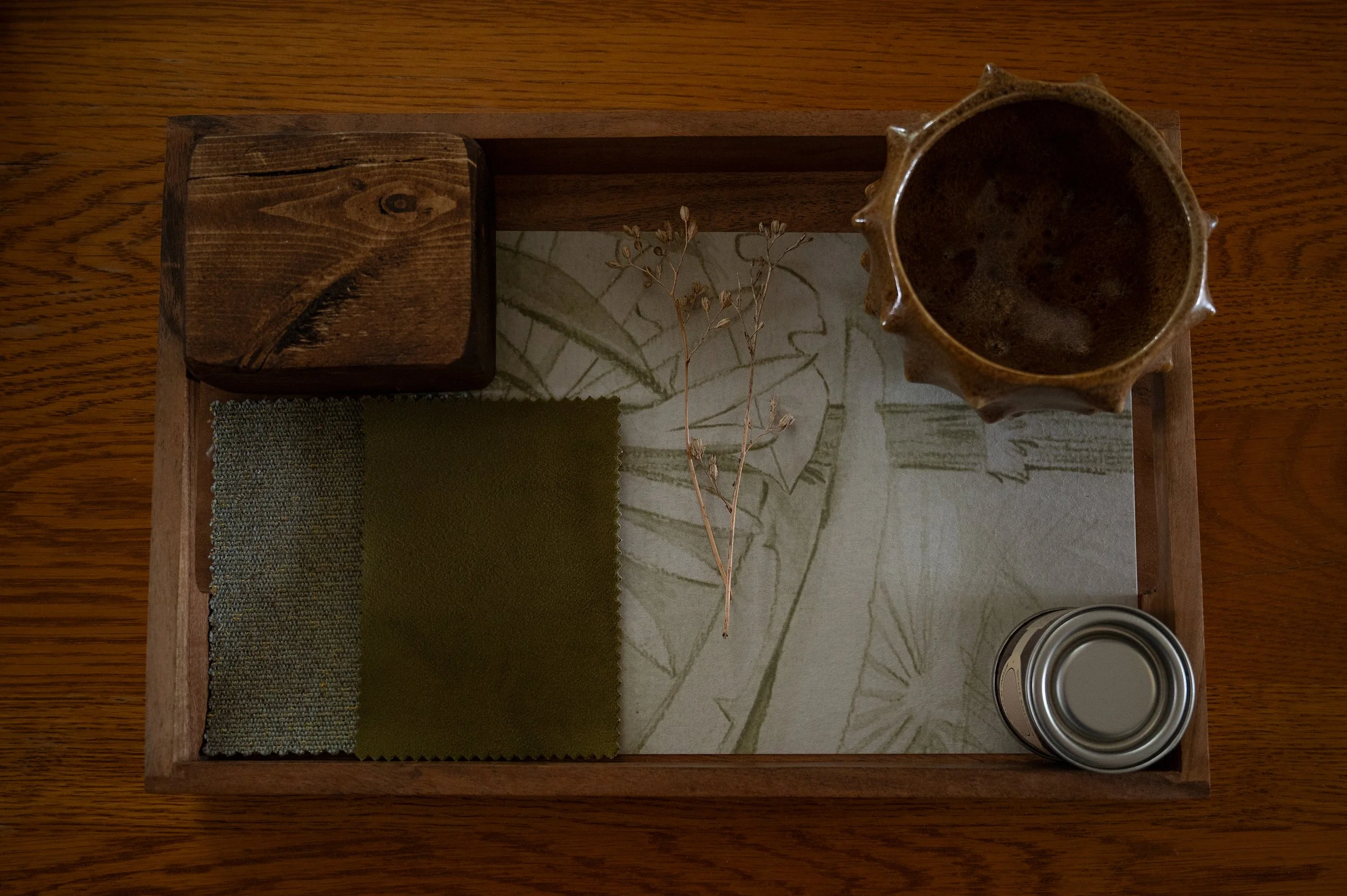 A wooden tray containing fabric swatches, a dried plant, a ceramic mug, and a metal can, all arranged on a wooden surface.
