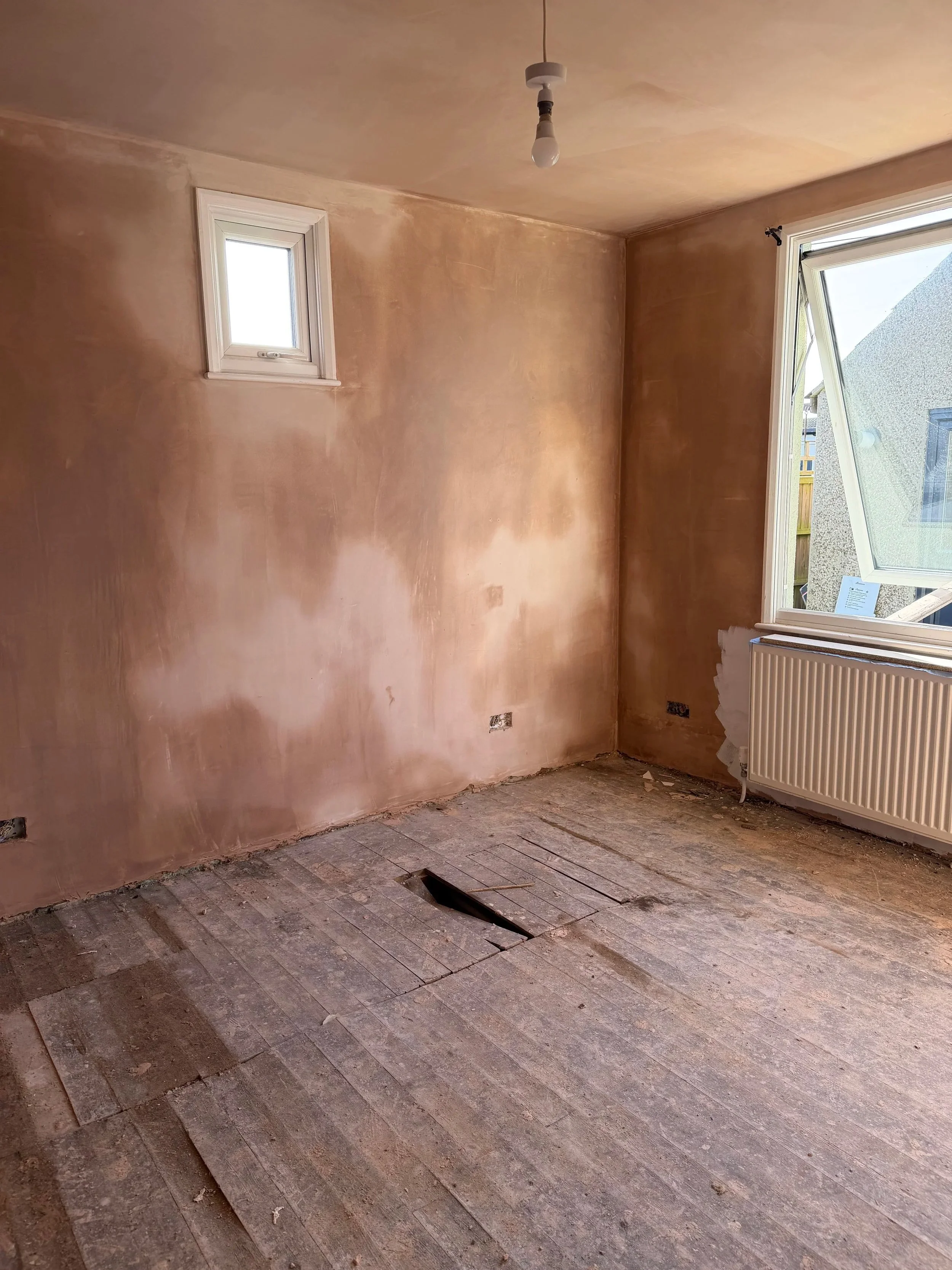 Room under renovation with an unfinished wooden floor, a wall with two windows, and a radiator. The floor has an open hole likely for flooring installation.