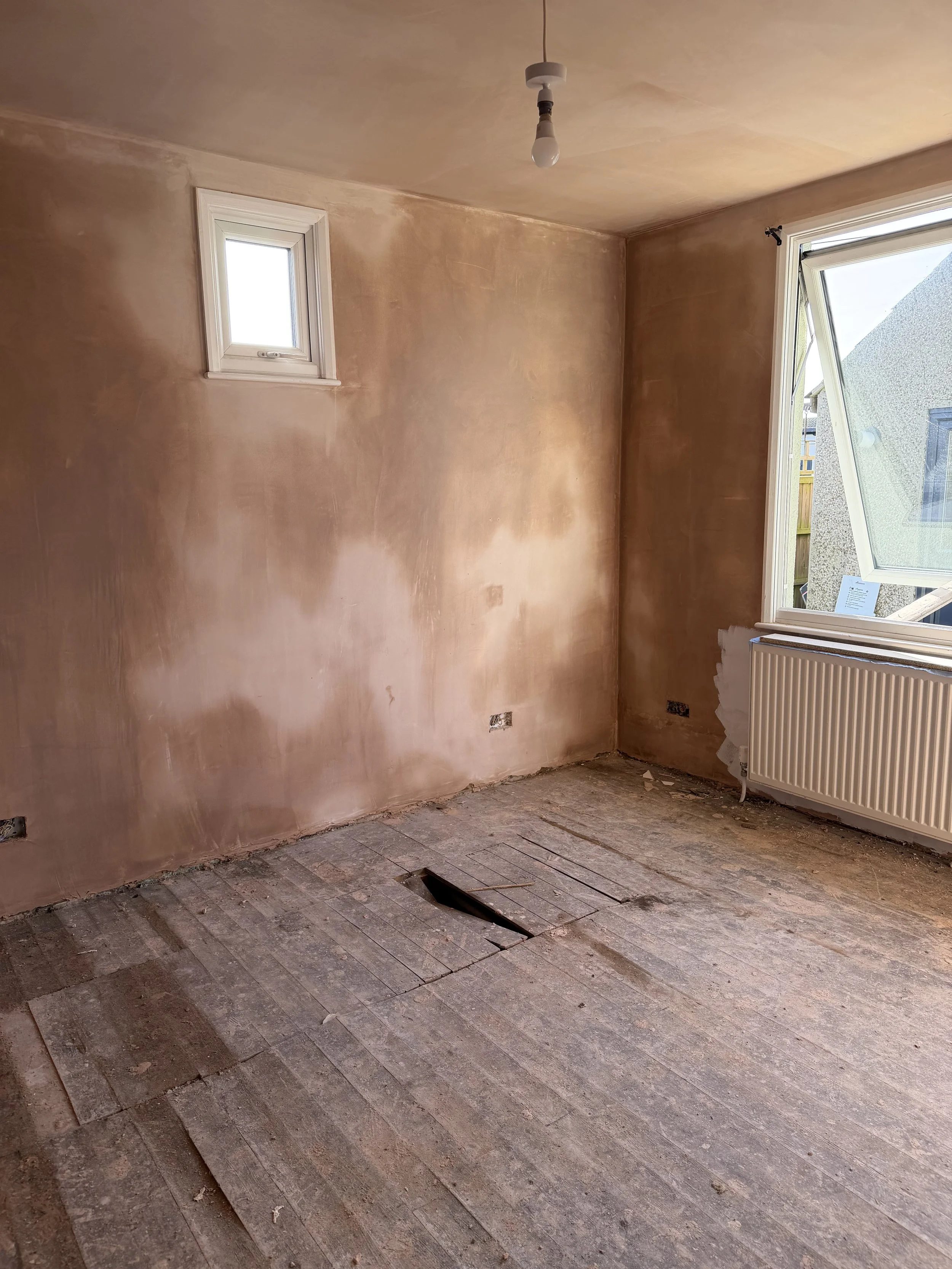 An empty room under renovation with bare wooden floors, unfinished walls, a small window at the top left, a larger window on the right with a view outside, and a radiator below the larger window. The ceiling has a single exposed lightbulb.