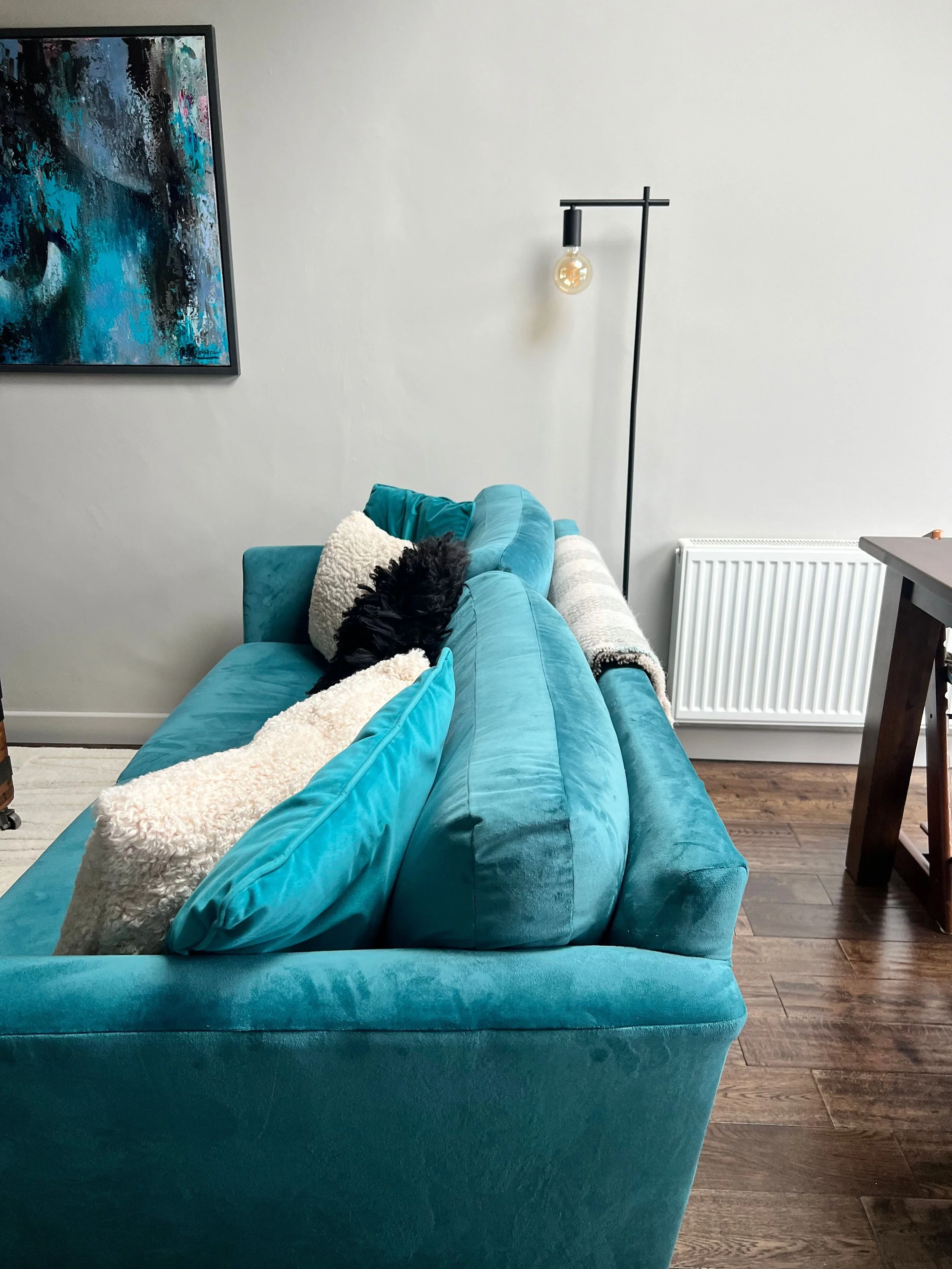 Living room with a turquoise velvet sofa, black and white pillows, a white textured pillow, a grey throw blanket, a modern floor lamp with an exposed bulb, artwork on the wall, wooden flooring, and a desk on the right.