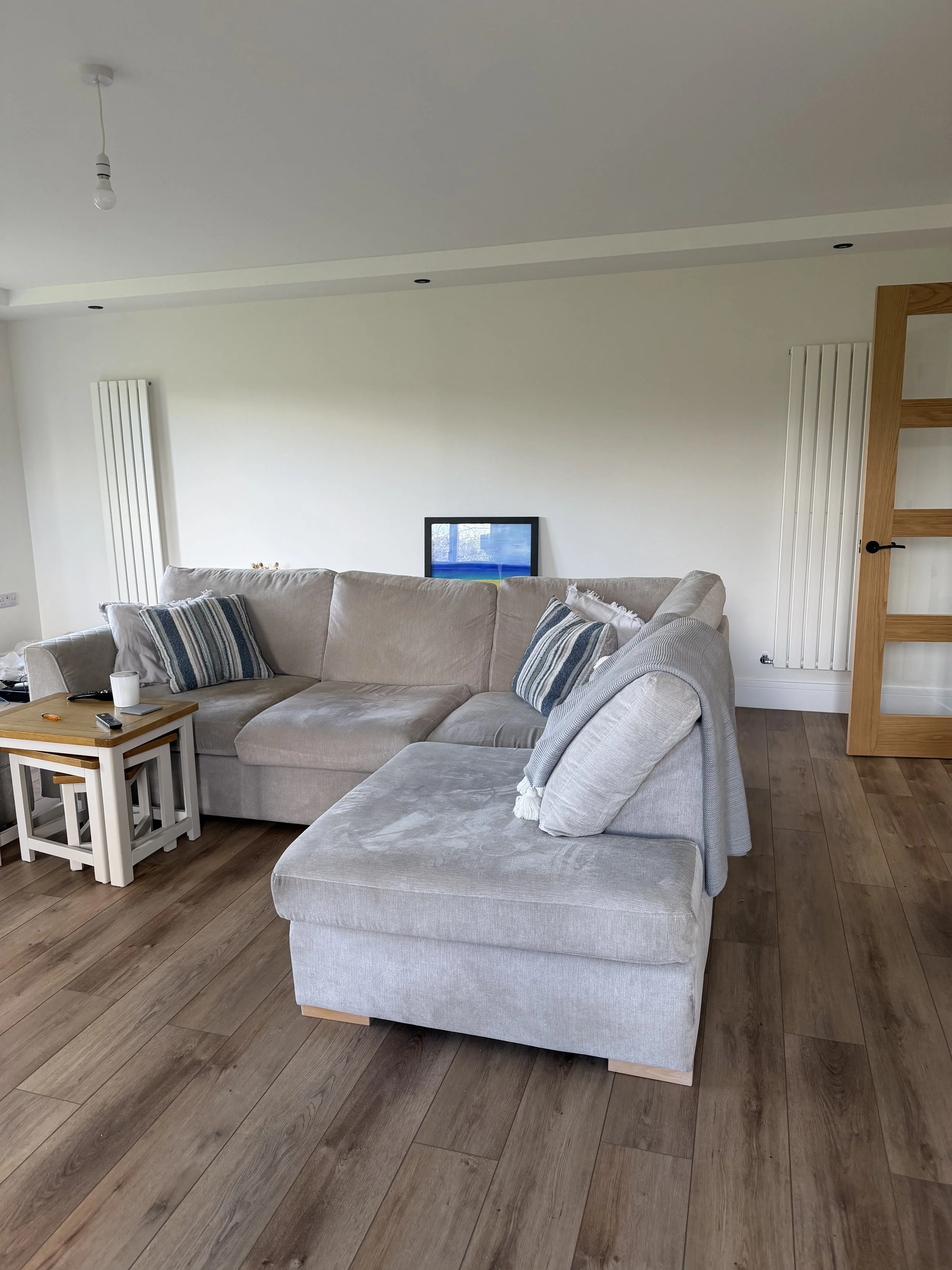 Living room with beige sectional sofa, striped pillows, small white side tables, a coffee mug, a TV on the wall, and wooden flooring.