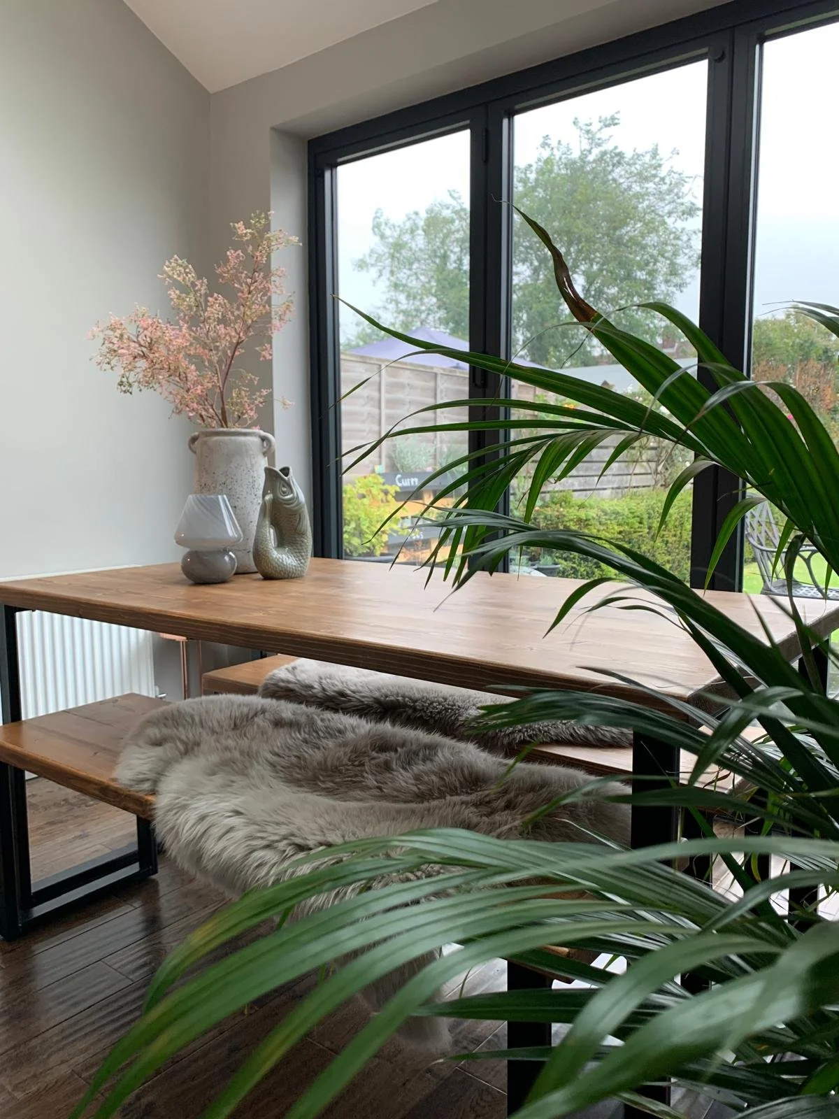 A cozy interior with a wooden table near large window, decorated with a pink flowering plant in a ceramic vase, a small white lamp, and a ceramic animal figurine. Fur throws and lush green plants are also on the table, with a view of a garden outside