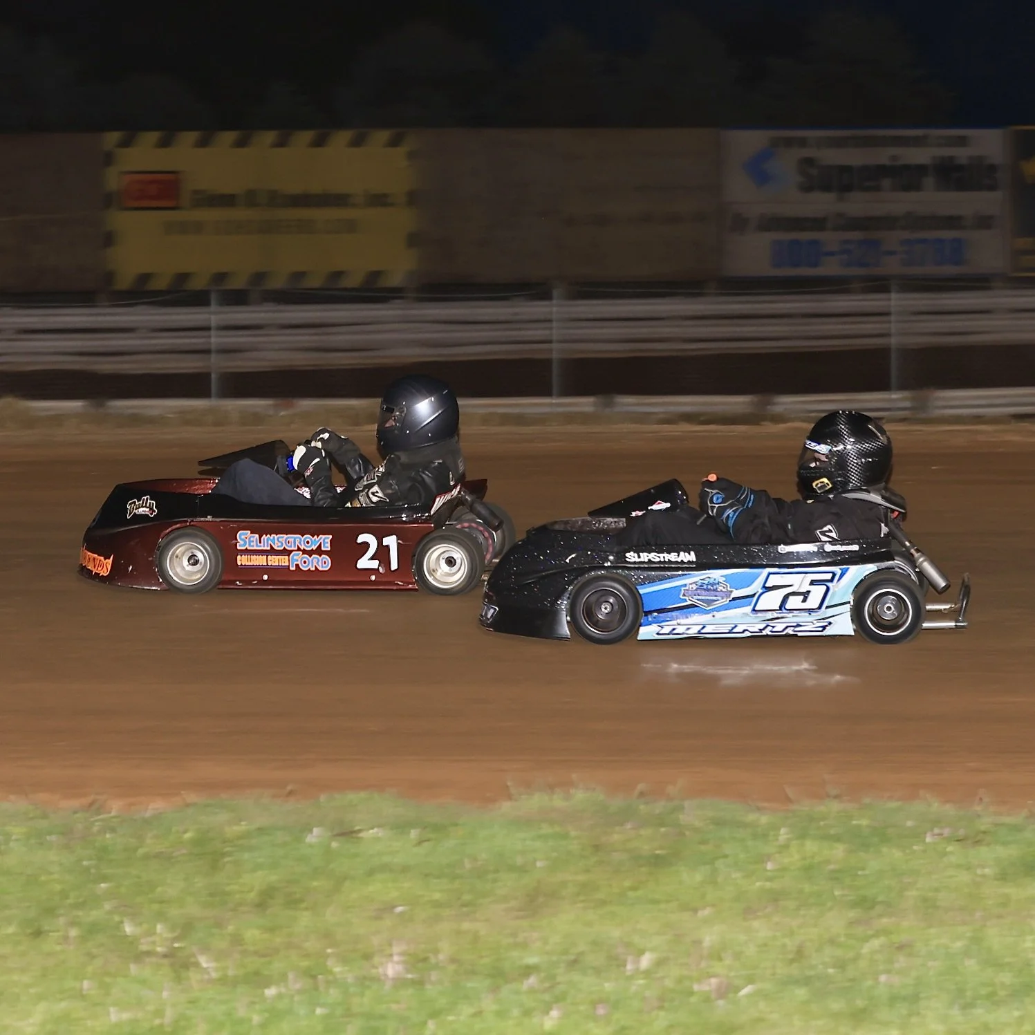 Two go-kart racers speeding on a dirt track, one in a red kart with number 21 and the other in a blue kart with number 25.
