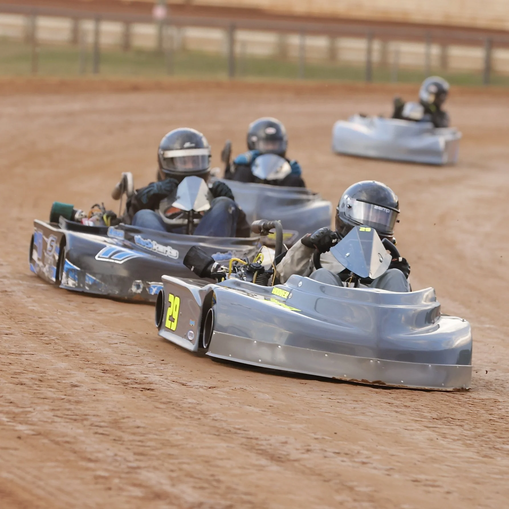 Group of go-kart racers competing on a dirt track, wearing helmets and racing suits.