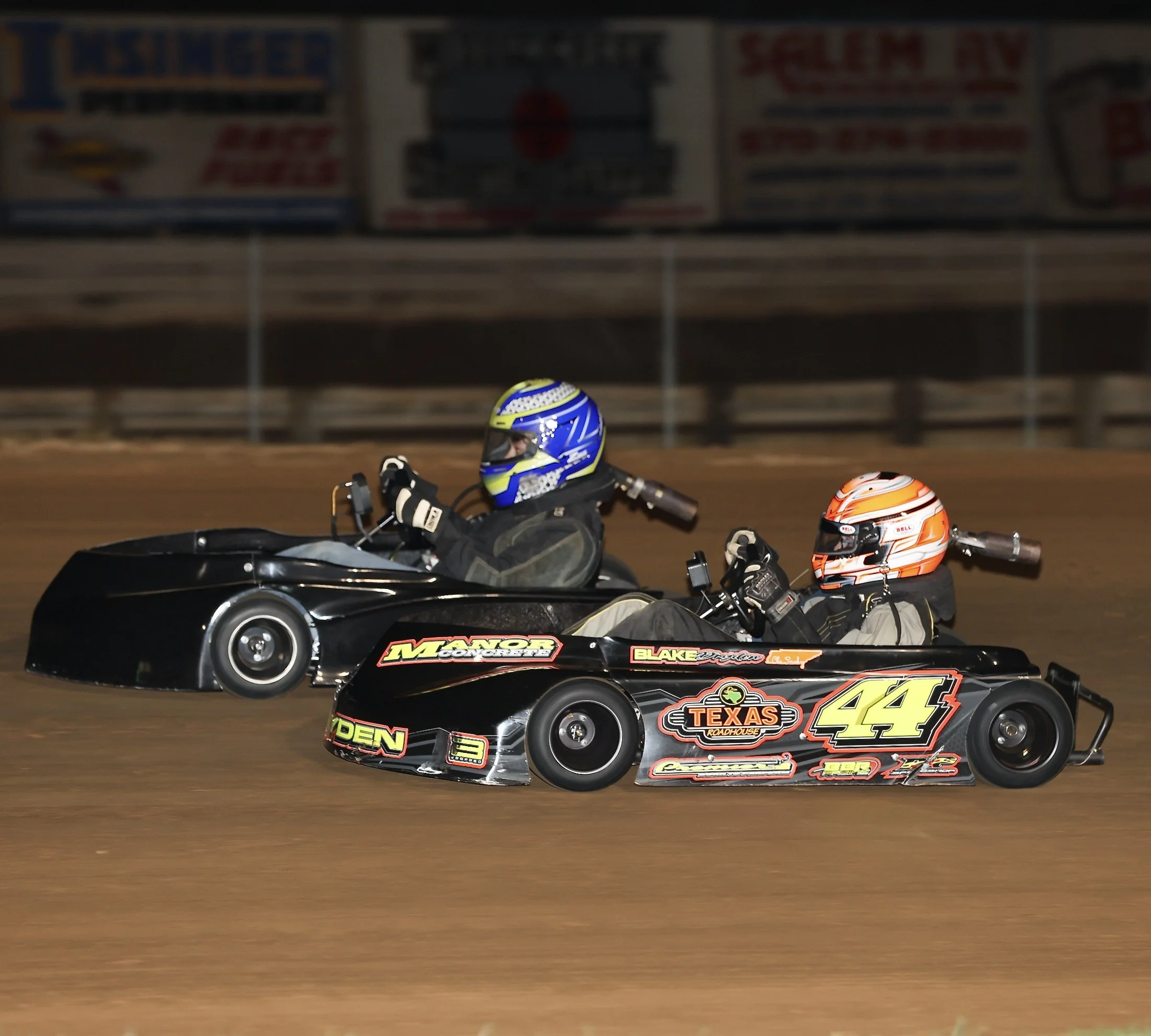 Two go-karts racing on a dirt track at night, driven by racers wearing helmets and protective gear. The karts have promotional logos and stickers.