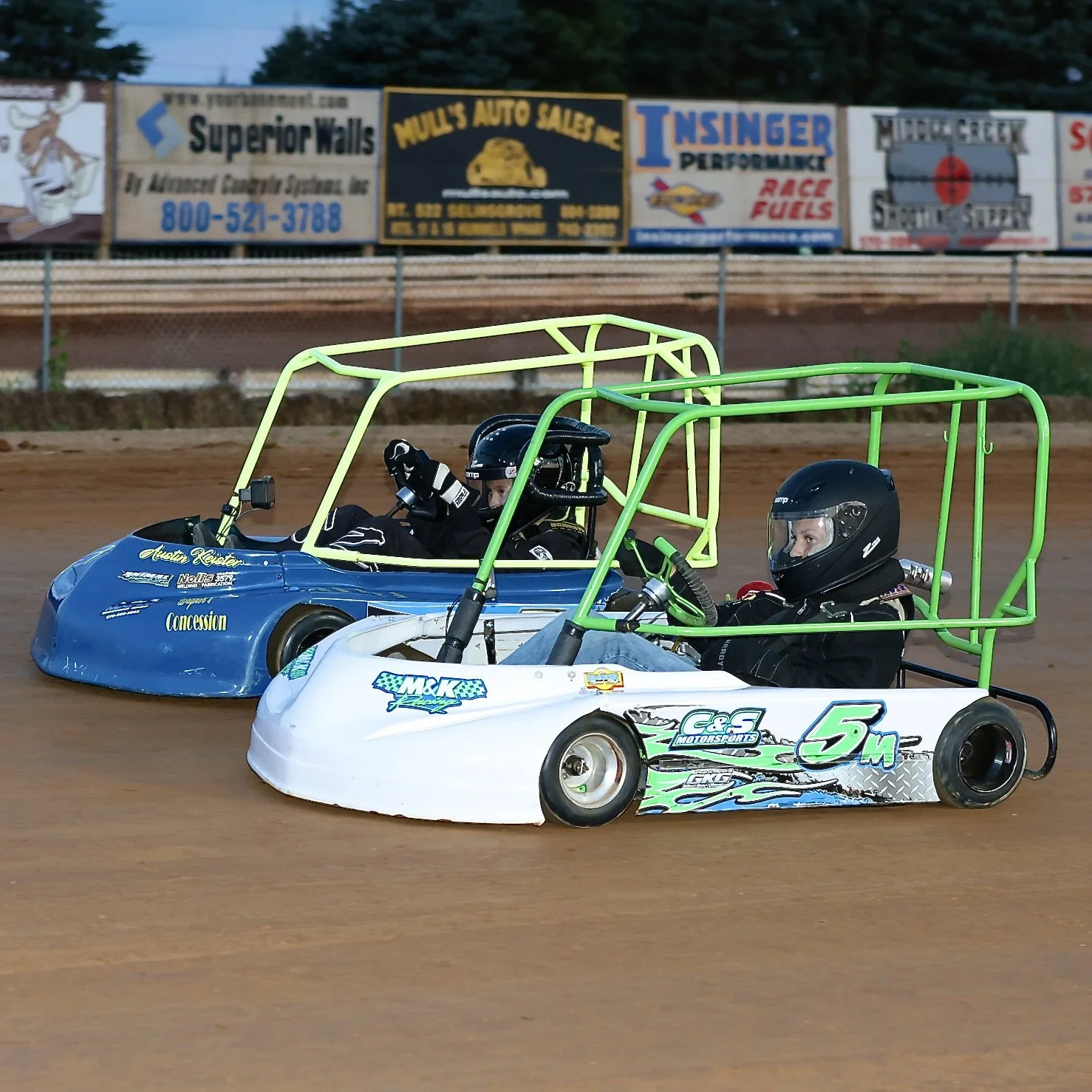 Two individuals in go-karts racing on a dirt track, wearing helmets and racing suits.