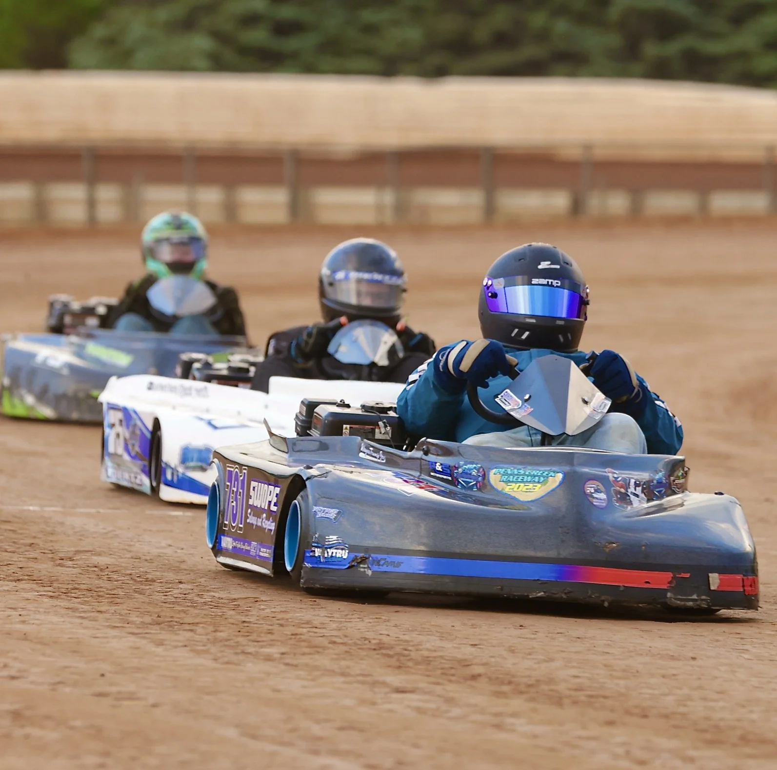 Go-kart racers on a dirt track, wearing helmets and racing gear.