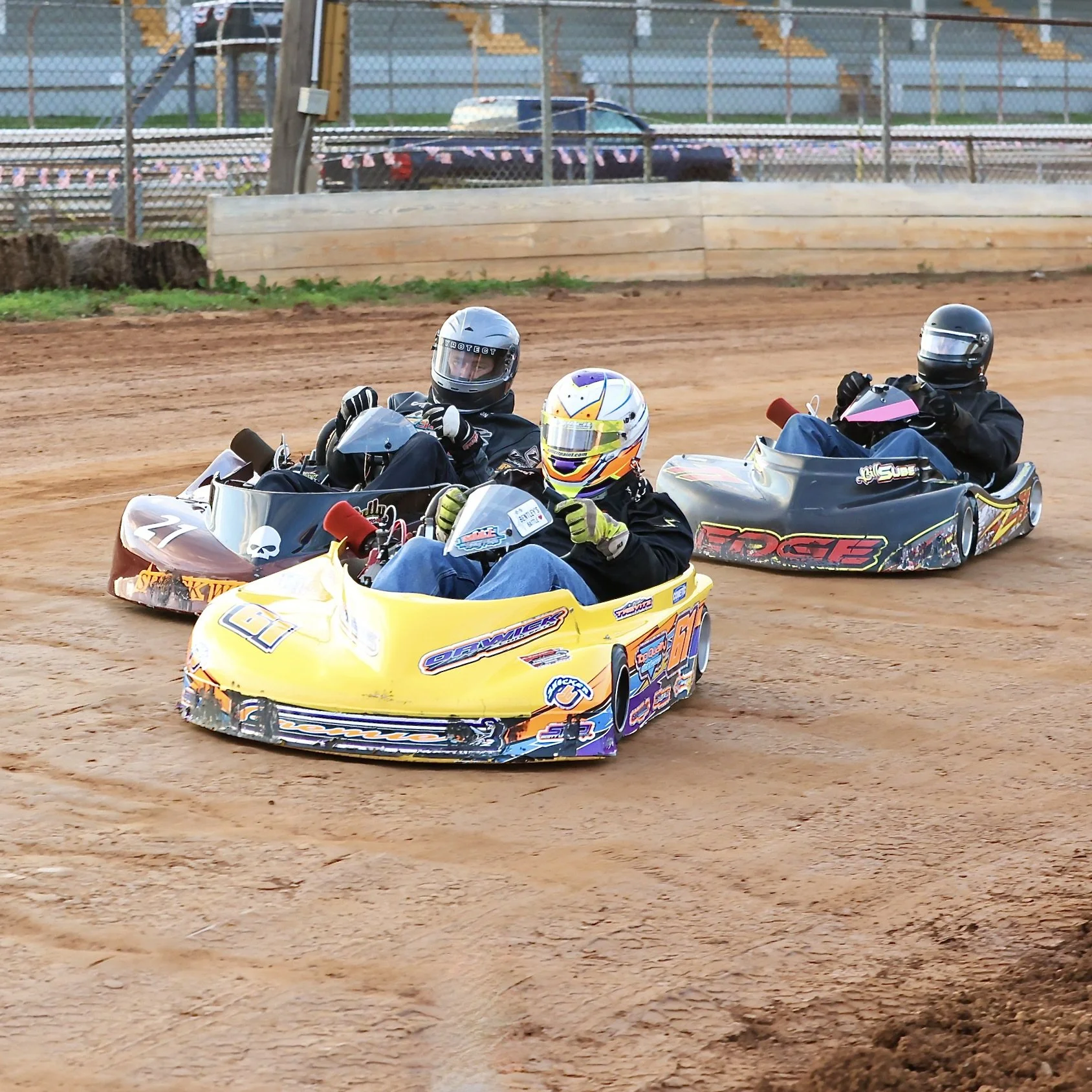 Three go-kart drivers racing on a dirt track, wearing helmets and protective gear. The lead kart is yellow, followed by two karts in different colors. Spectator stands and barriers are visible in the background.