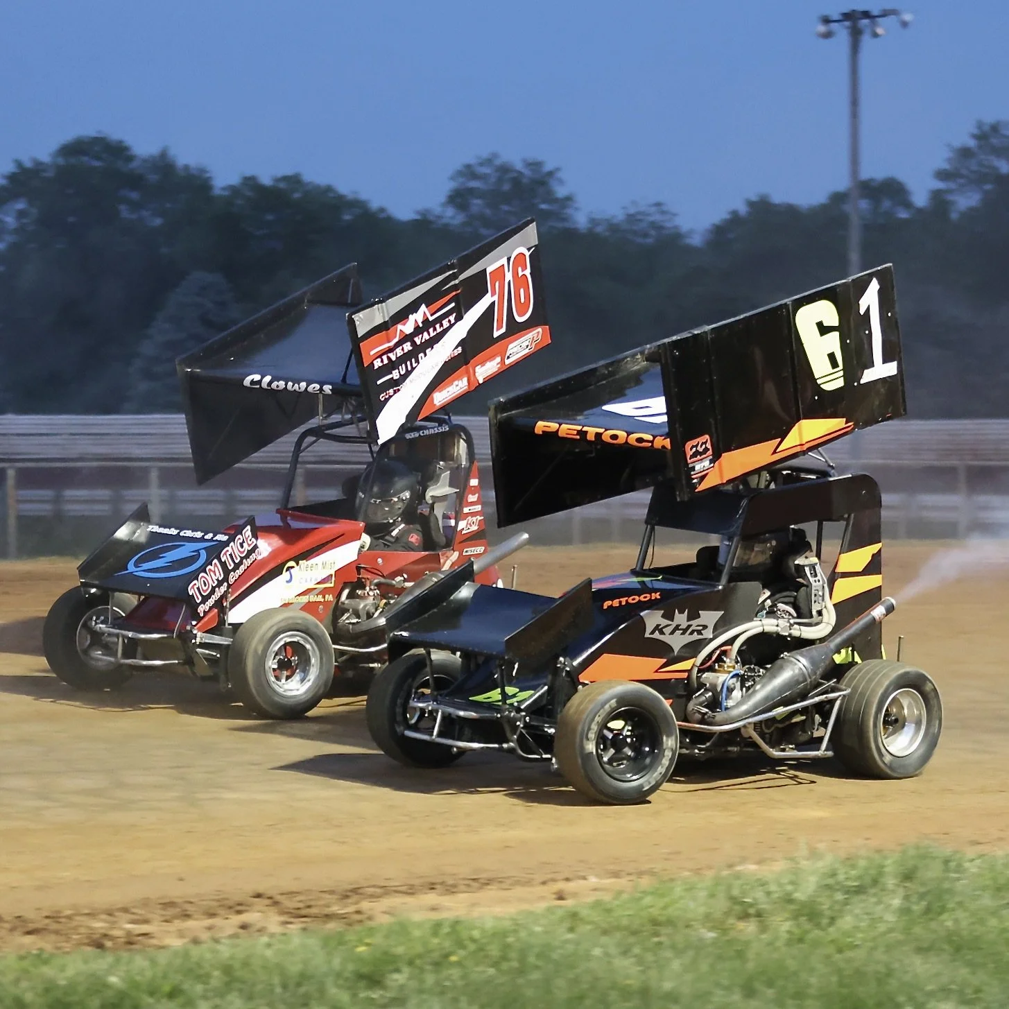 Two sprint cars racing on a dirt track at dusk.