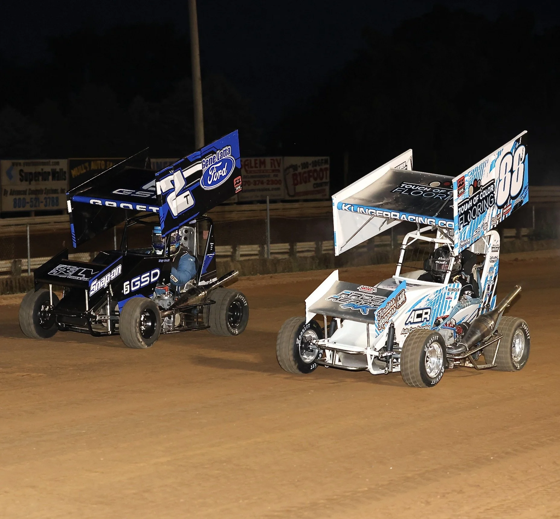 Two sprint cars racing on a dirt track at night, each with colorful liveries and large rear wings.