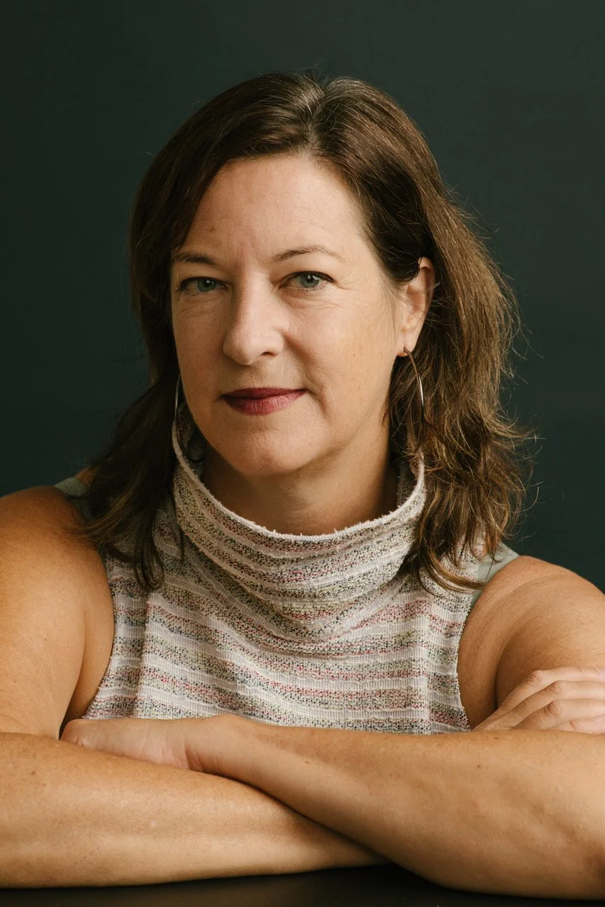 A middle-aged woman with shoulder-length brown hair and light skin, wearing a sleeveless, high-neck, multicolored fabric top, sitting with her arms crossed on a dark surface against a dark background.