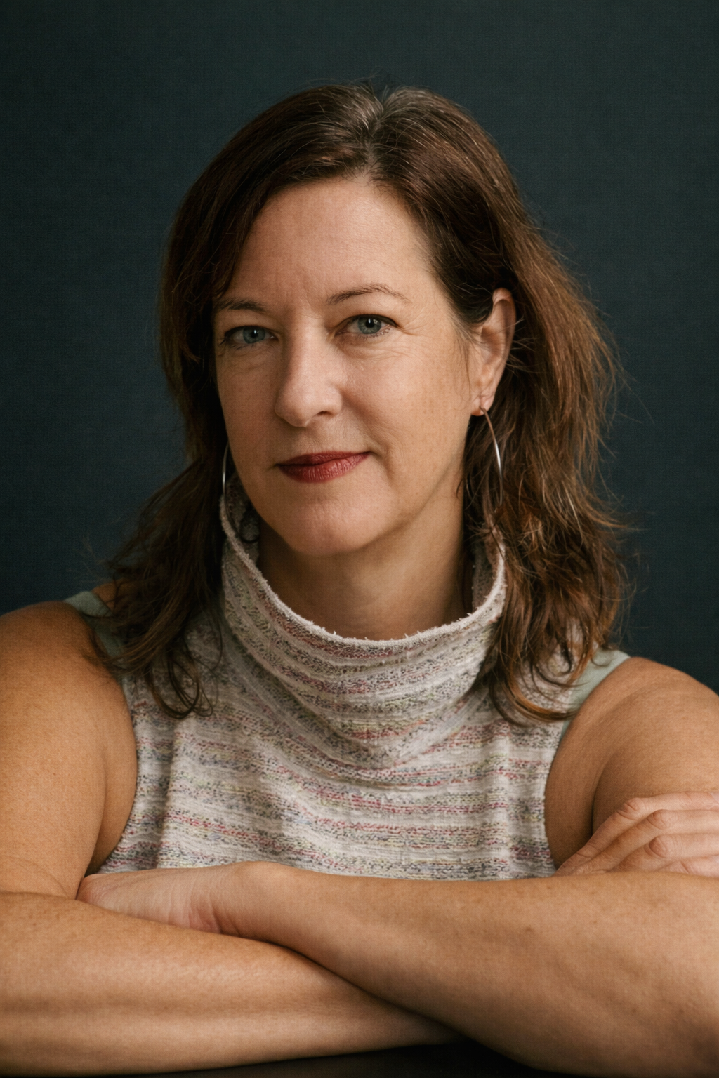 A middle-aged woman with shoulder-length brown hair and light skin, wearing a sleeveless, high-neck, multicolored fabric top, sitting with her arms crossed on a dark surface against a dark background.