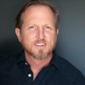 Portrait of a man with light skin, brown hair, and a beard, smiling and wearing a dark collared shirt against a dark background.