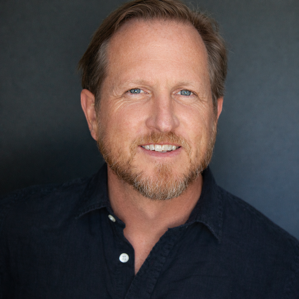 Portrait of a man with light skin, brown hair, and a beard, smiling and wearing a dark collared shirt against a dark background.
