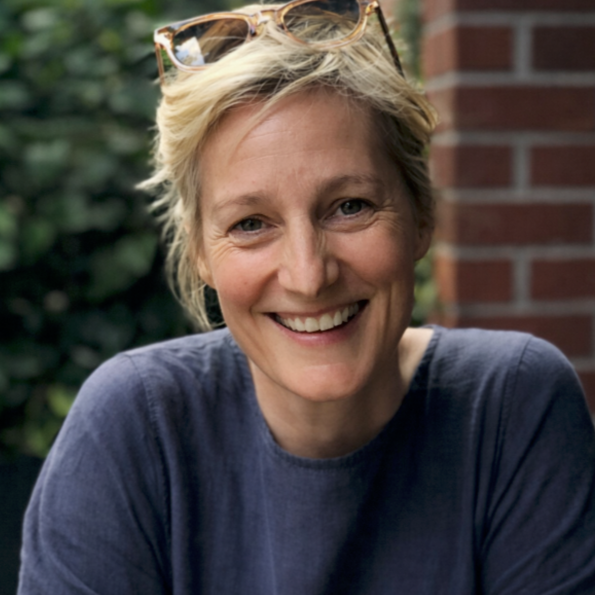 Smile of a young woman with curly hair, glasses, and jewelry, in a cozy indoor setting.