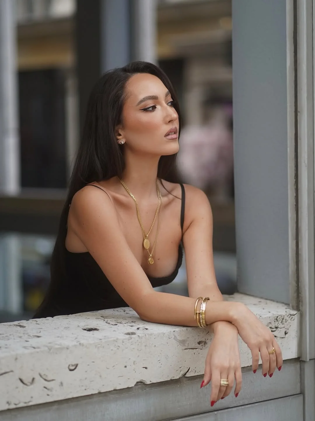 A woman with long dark hair and makeup, wearing a black spaghetti strap top, gold jewelry, and rings, is looking out from a window ledge with a thoughtful expression.