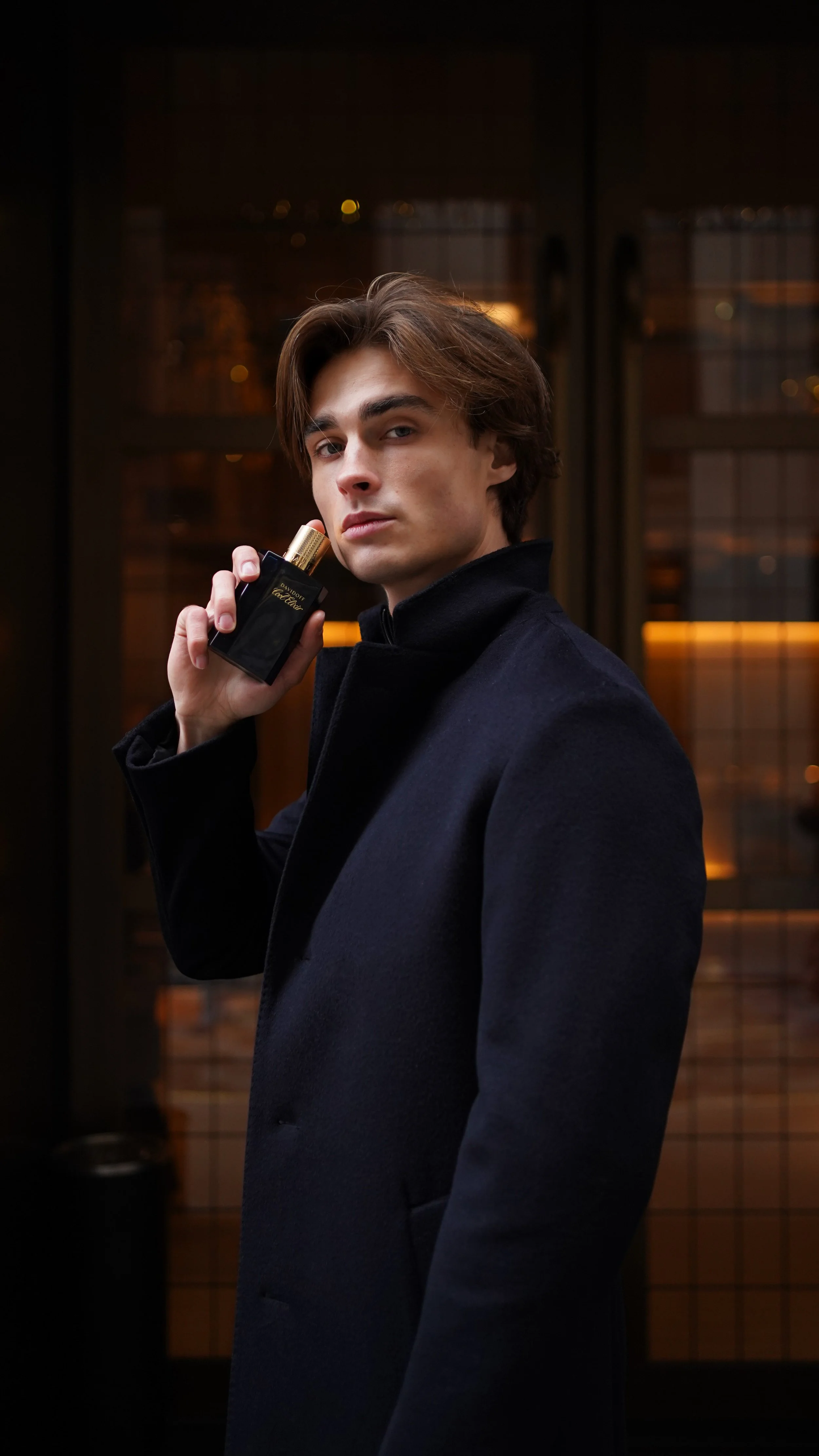 A young man in a dark coat holding a black perfume bottle with a gold cap, standing in front of a window with grid-like panes reflecting warm interior lighting.