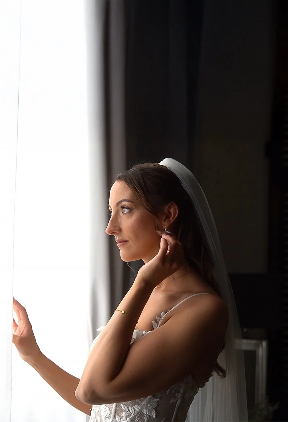 Jeune femme en robe de mariée ajustant une boucle d'oreille, près d'une fenêtre avec rideaux, en intérieur