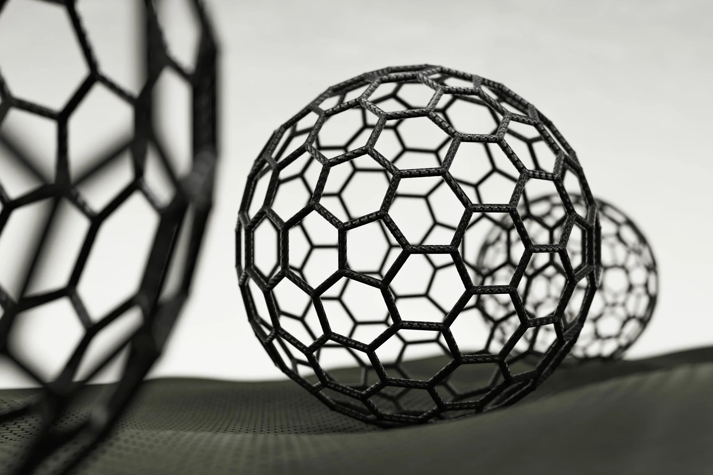 Close-up of a black geometric wireframe sculpture resembling a sphere, with other similar shapes blurred in the background, set against a plain light-colored background.