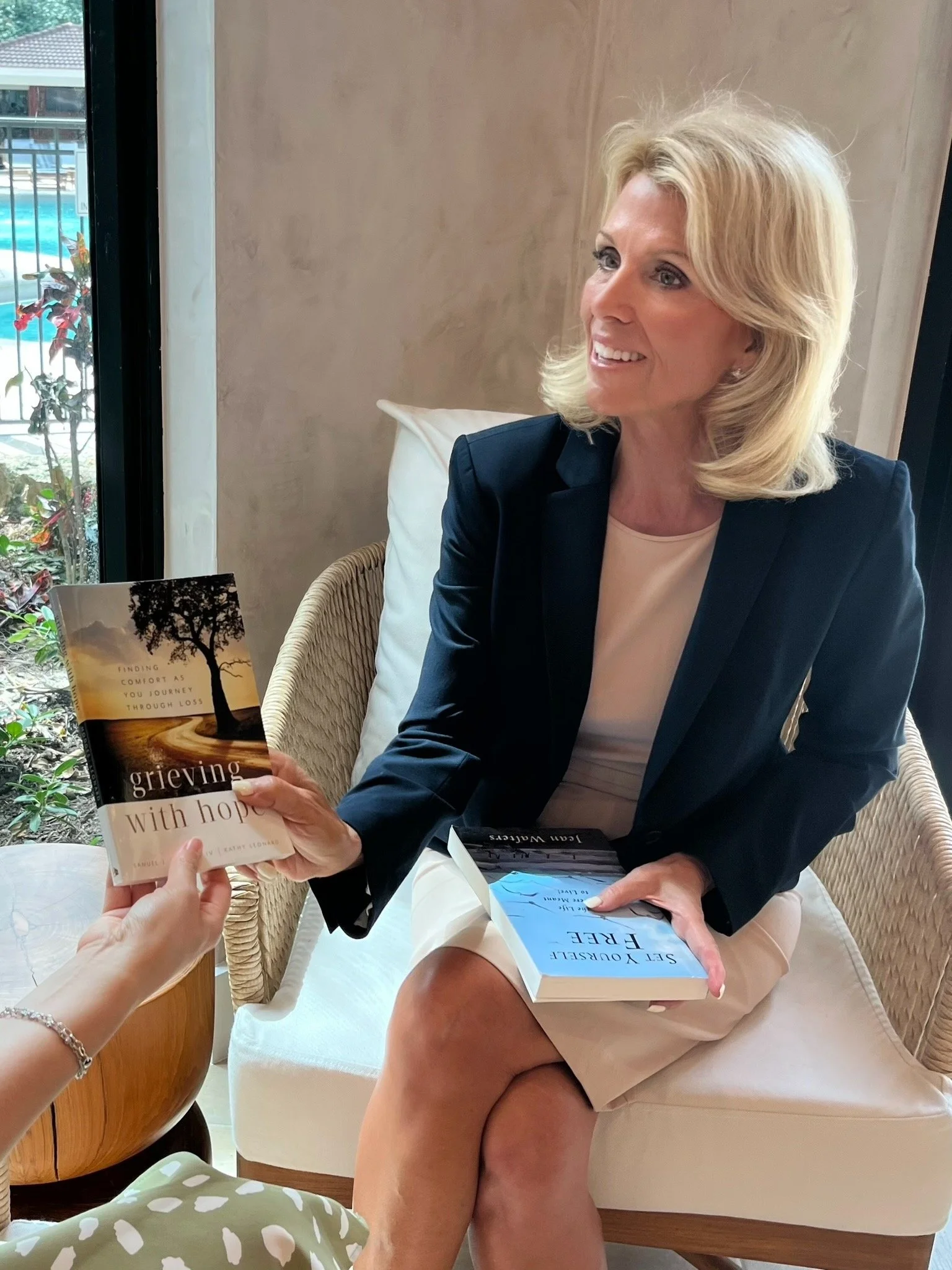 A woman with blonde hair wearing a black blazer and beige dress sitting on a white cushioned chair. She is smiling and receiving a book titled 'Grieving with Hope' from another person. The background includes a window showing part of a pool and outdoor area.