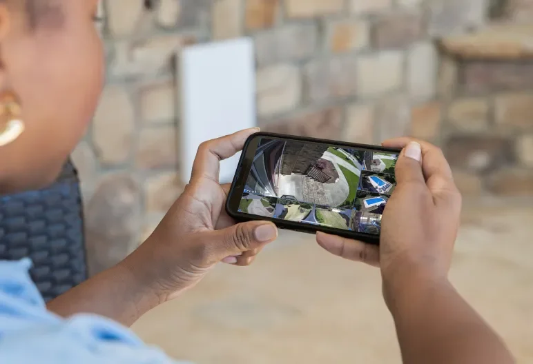 Person holding a smartphone taking a photo of a small outdoor area with grass, pathway, and outdoor furniture.