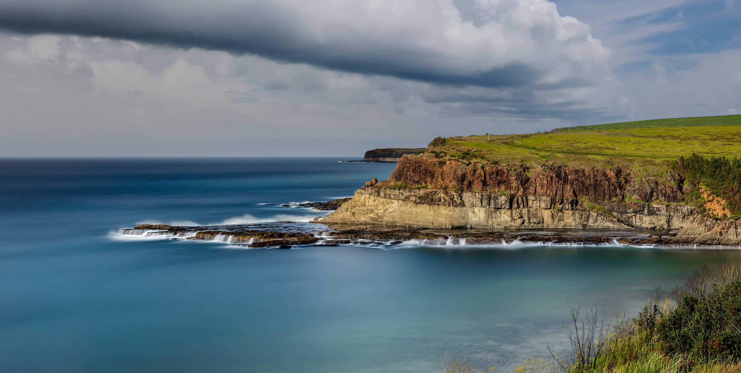 Coastal cliffs with layers of rock, green grass, and a cloudy sky above the ocean.