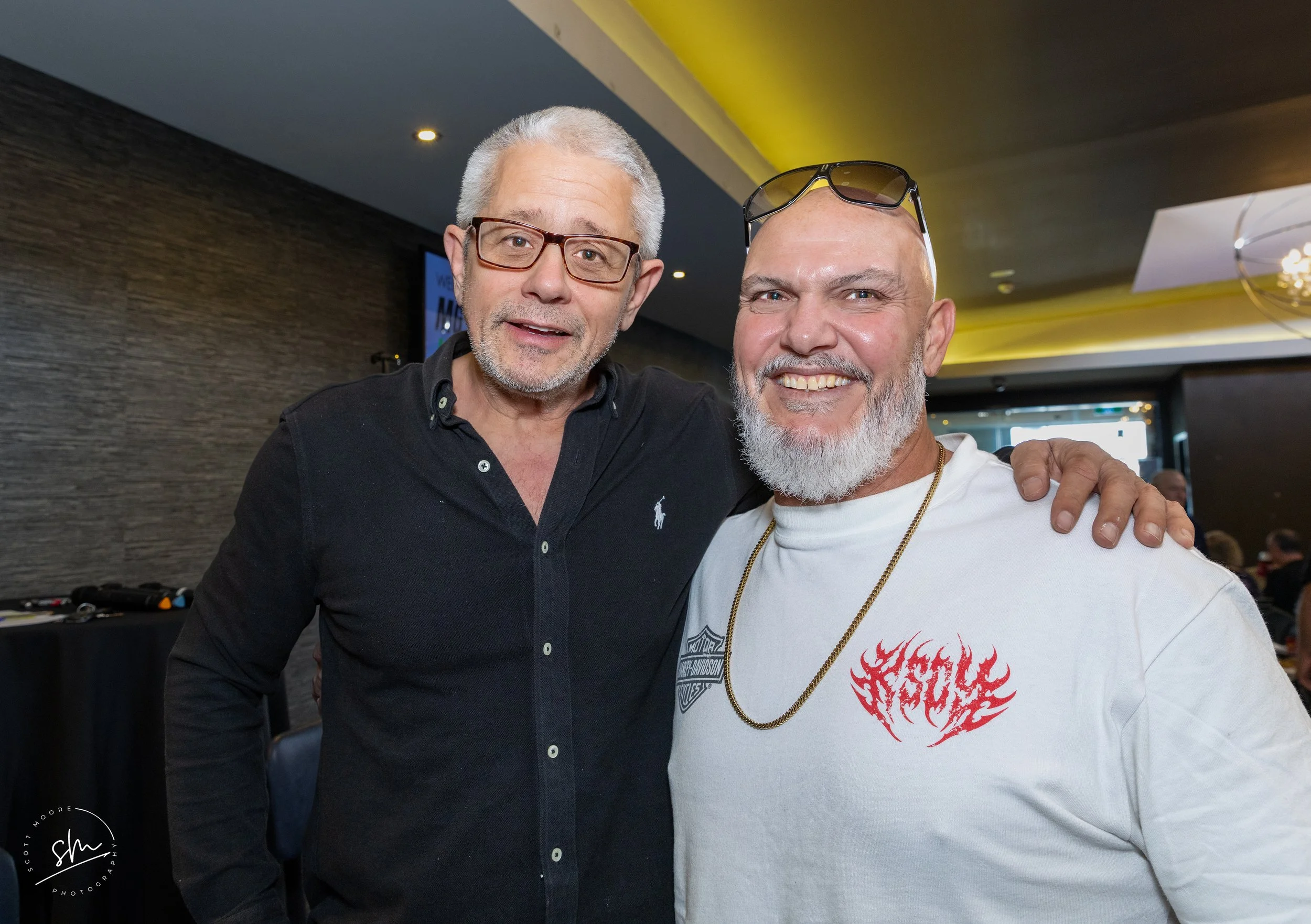 Two men smiling and posing together indoors, one wearing glasses and a black shirt, the other wearing sunglasses on his head and a white t-shirt with a red flame design.