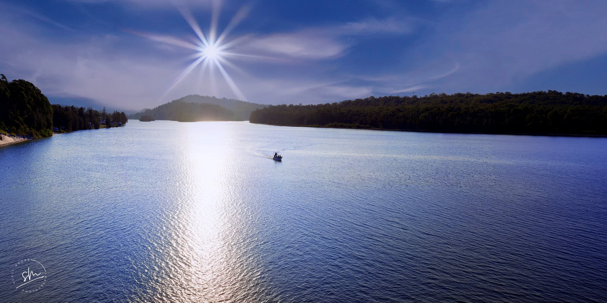 A boat with two people inside floating on a calm, reflective body of water under a bright sun in a clear blue sky. Forested hills are visible on both sides of the water.