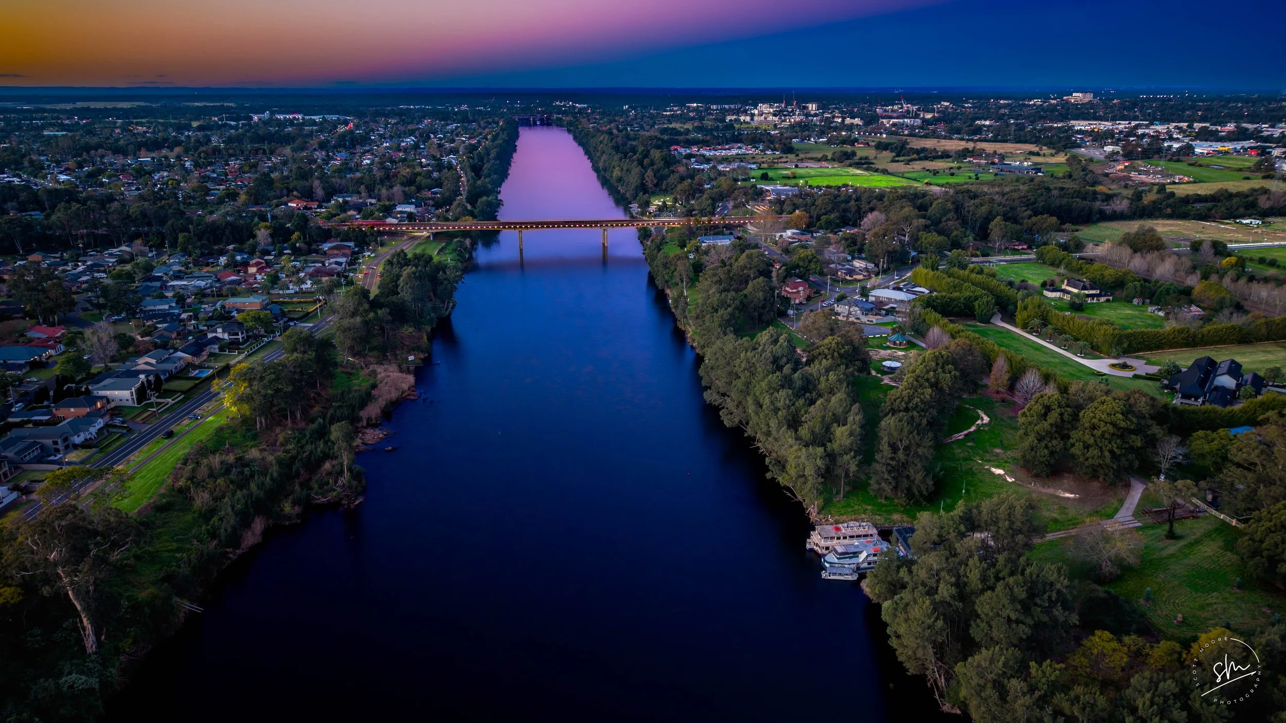 Aerial view of a river running through a town at dusk, with a bridge illuminated with lights crossing over the river, and a sky transitioning from pink to dark blue.