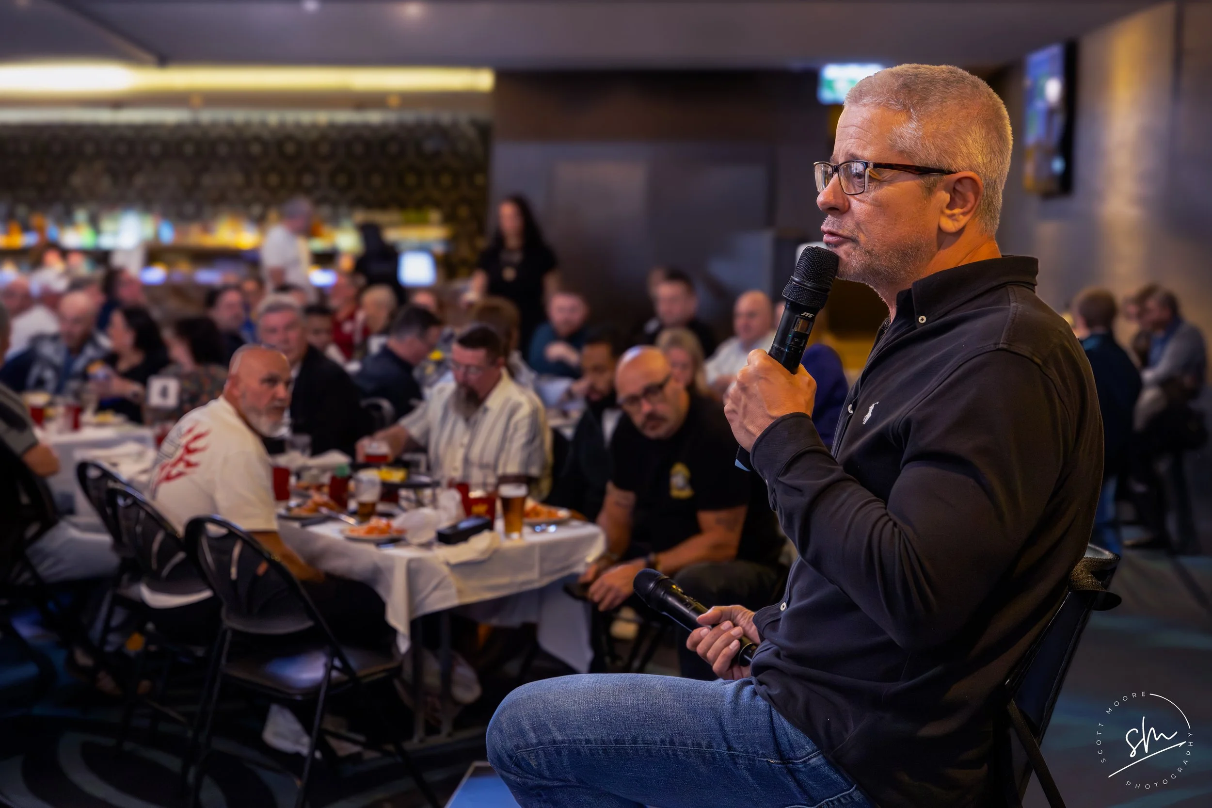 A man with glasses and gray hair speaking into a microphone at a crowded indoor event with many seated attendees eating at tables. anthony Maroon talking and hosting function