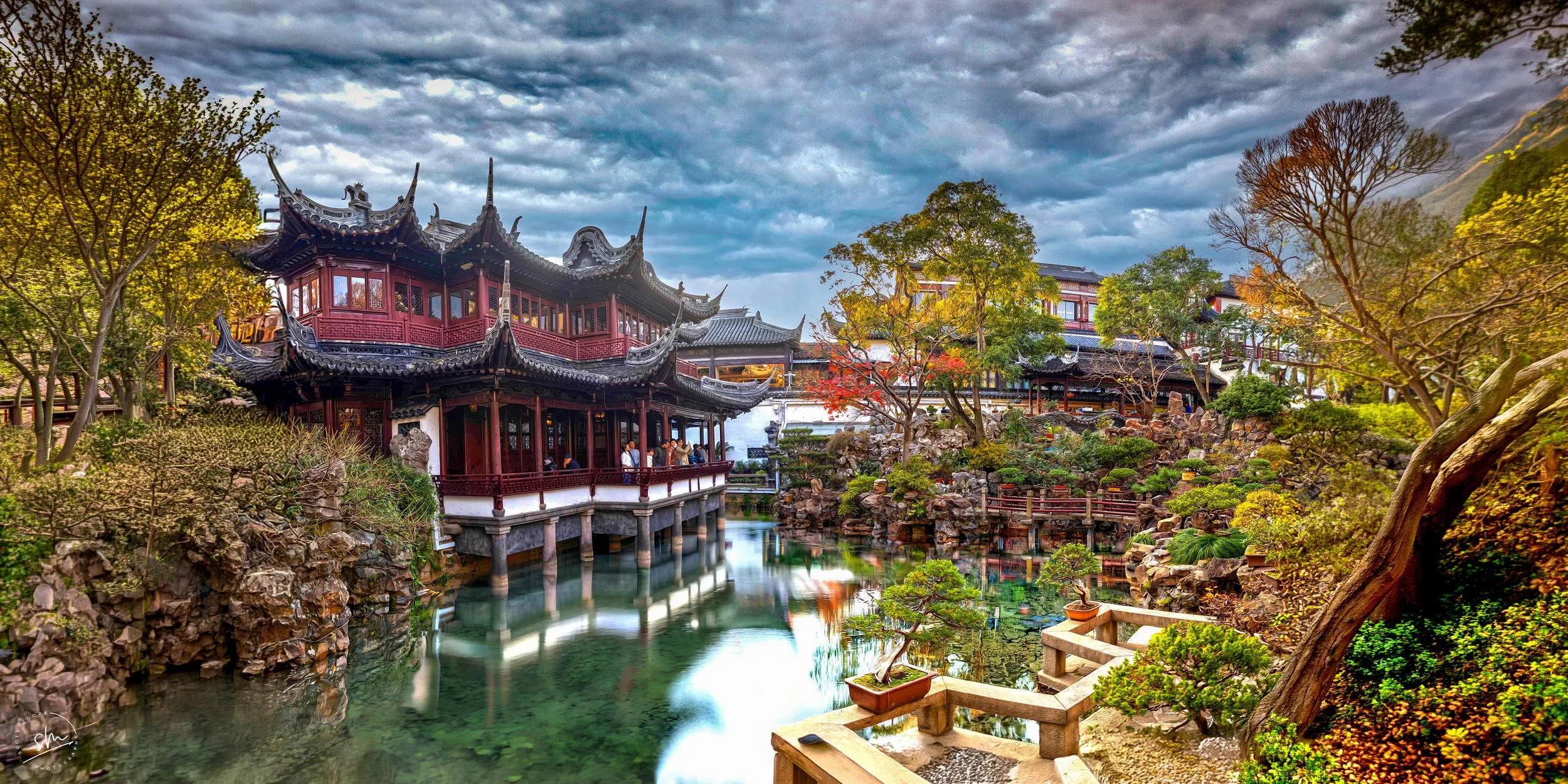 Traditional Chinese garden with pagoda-style building, pond, rocks, and lush trees under a cloudy sky.