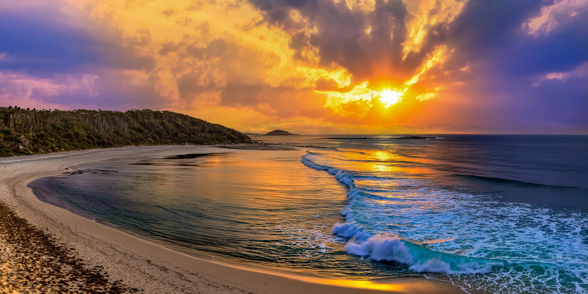 Sun setting over the ocean with colorful clouds, waves gently rolling onto a sandy beach, and a forested coastline in the distance.